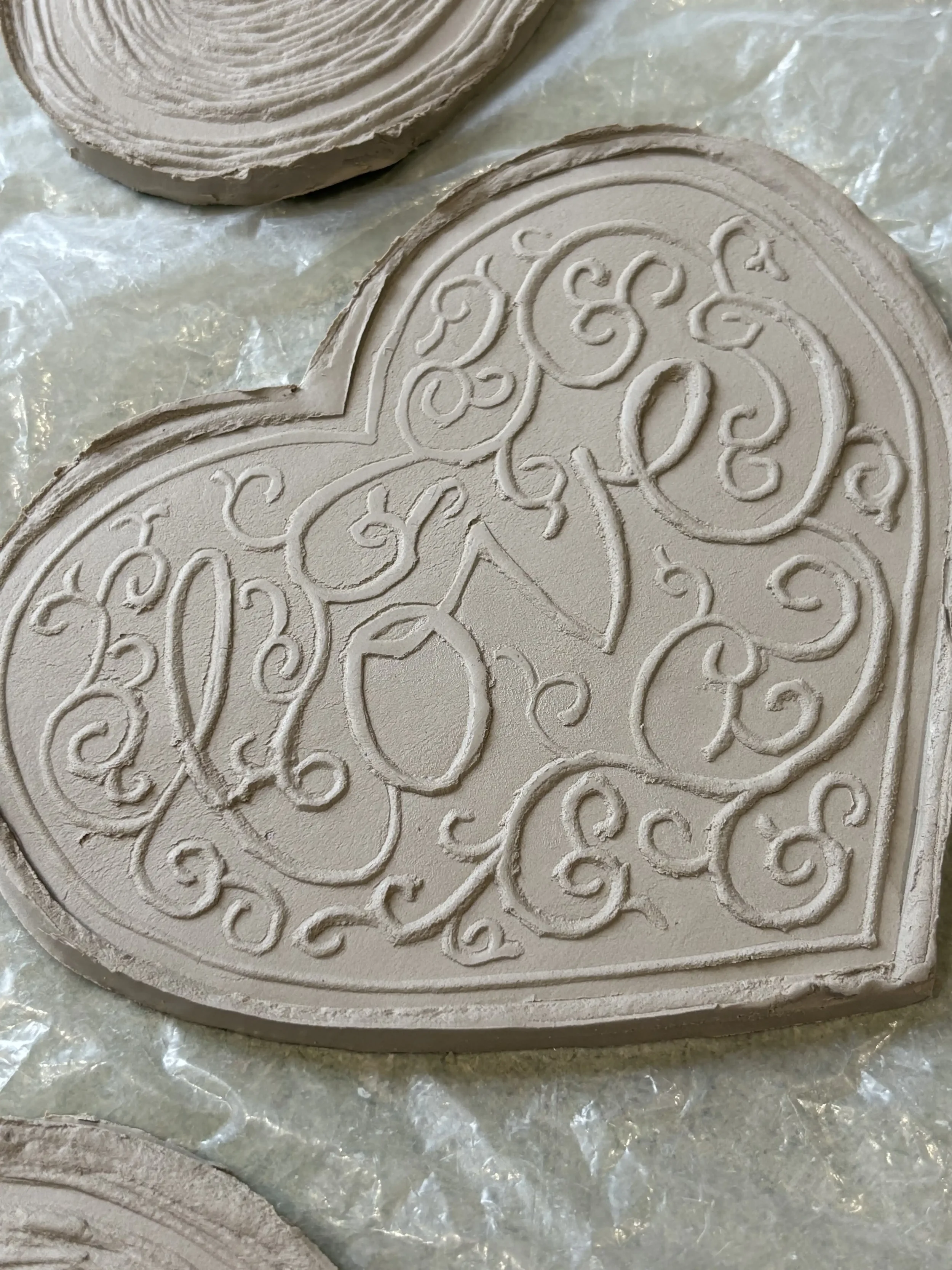 A heart-shaped textured clay tile with intricate swirl and cursive designs and the word 'love' etched into its surface.