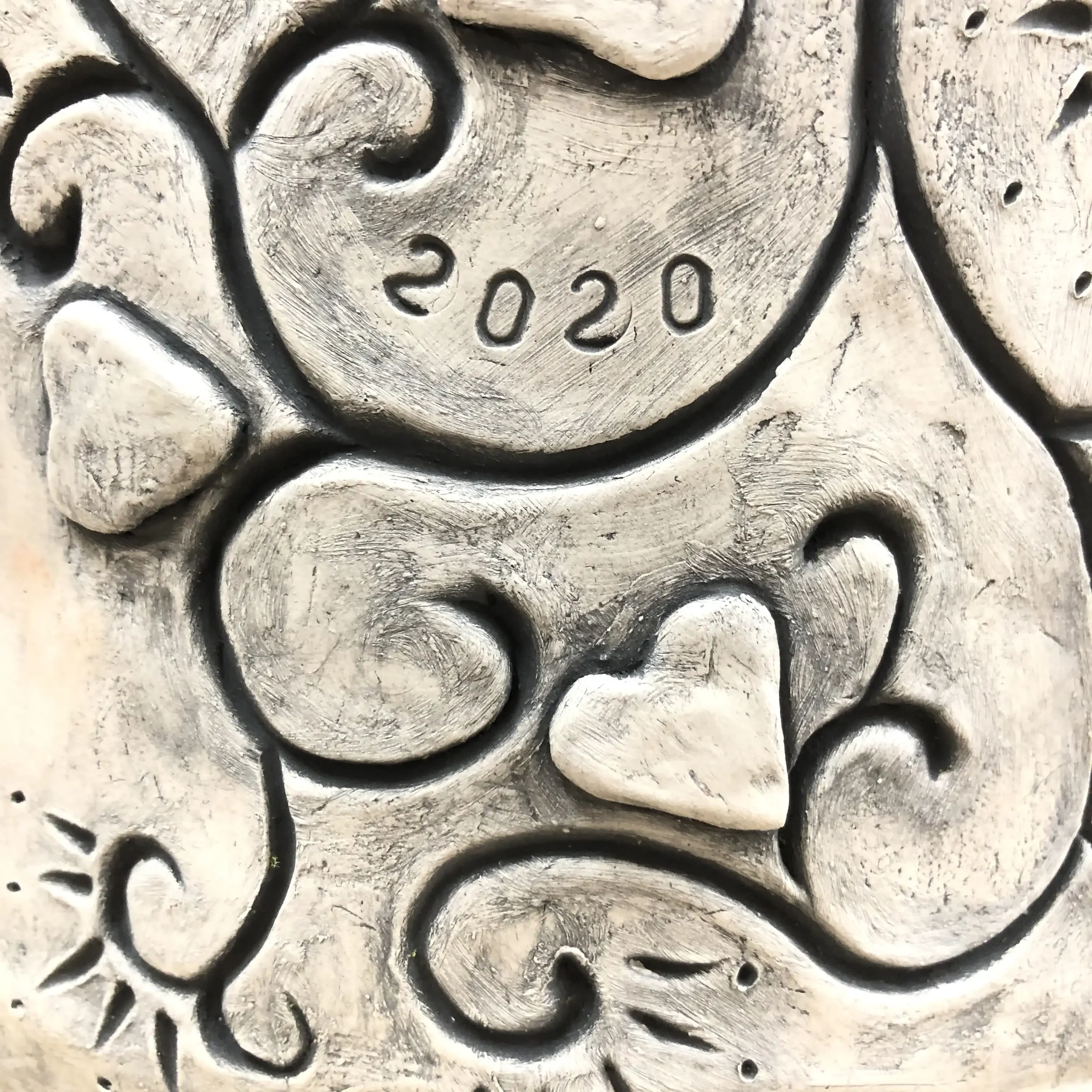 Close-up of an ornate clay pot surface with a heart shape near the centre, the year 2020 stamped in the upper area, and decorative scrollwork and patterns.