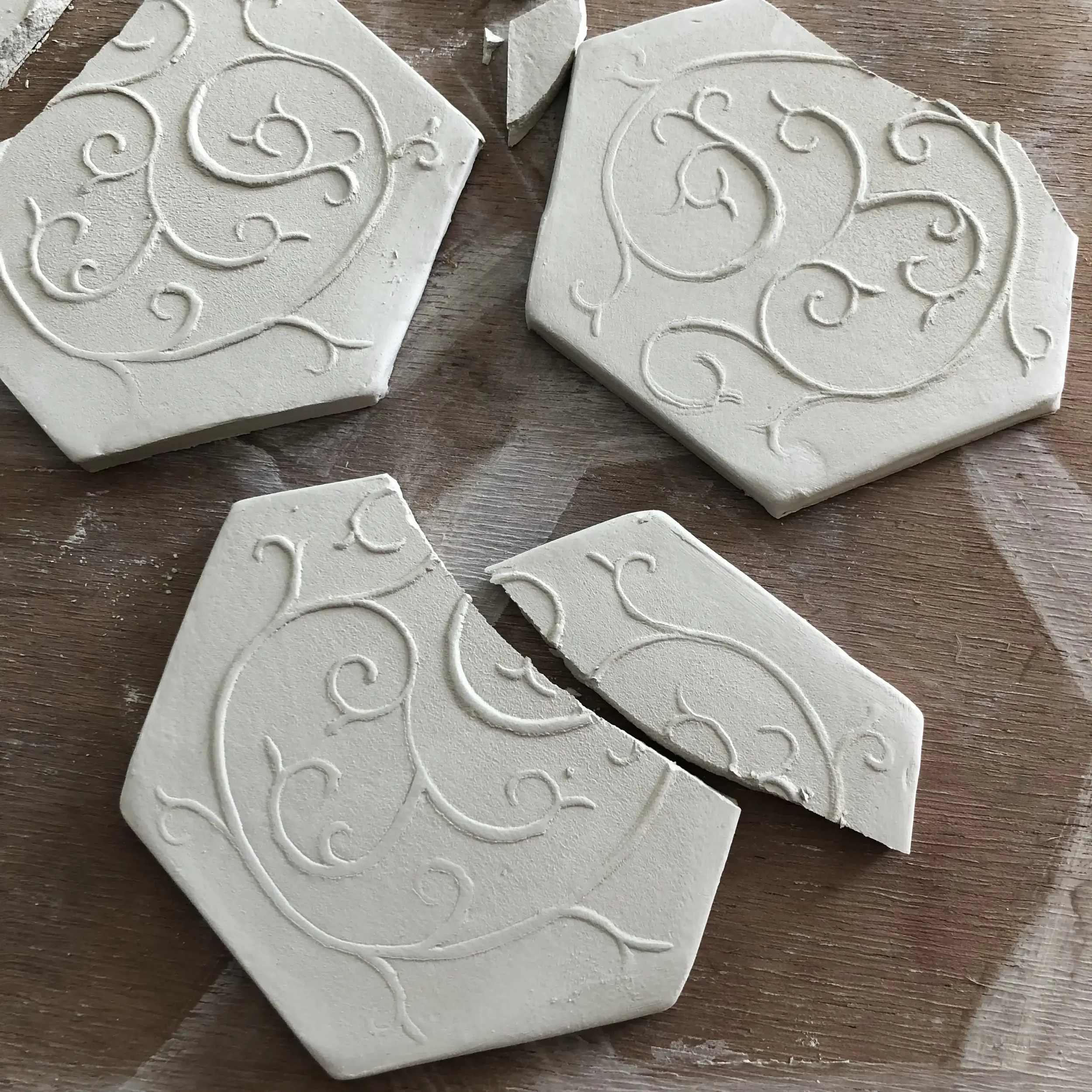 Decorative white hexagon shaped ceramic tiles with intricate swirling patterns, some broken into pieces, placed on a wooden surface.