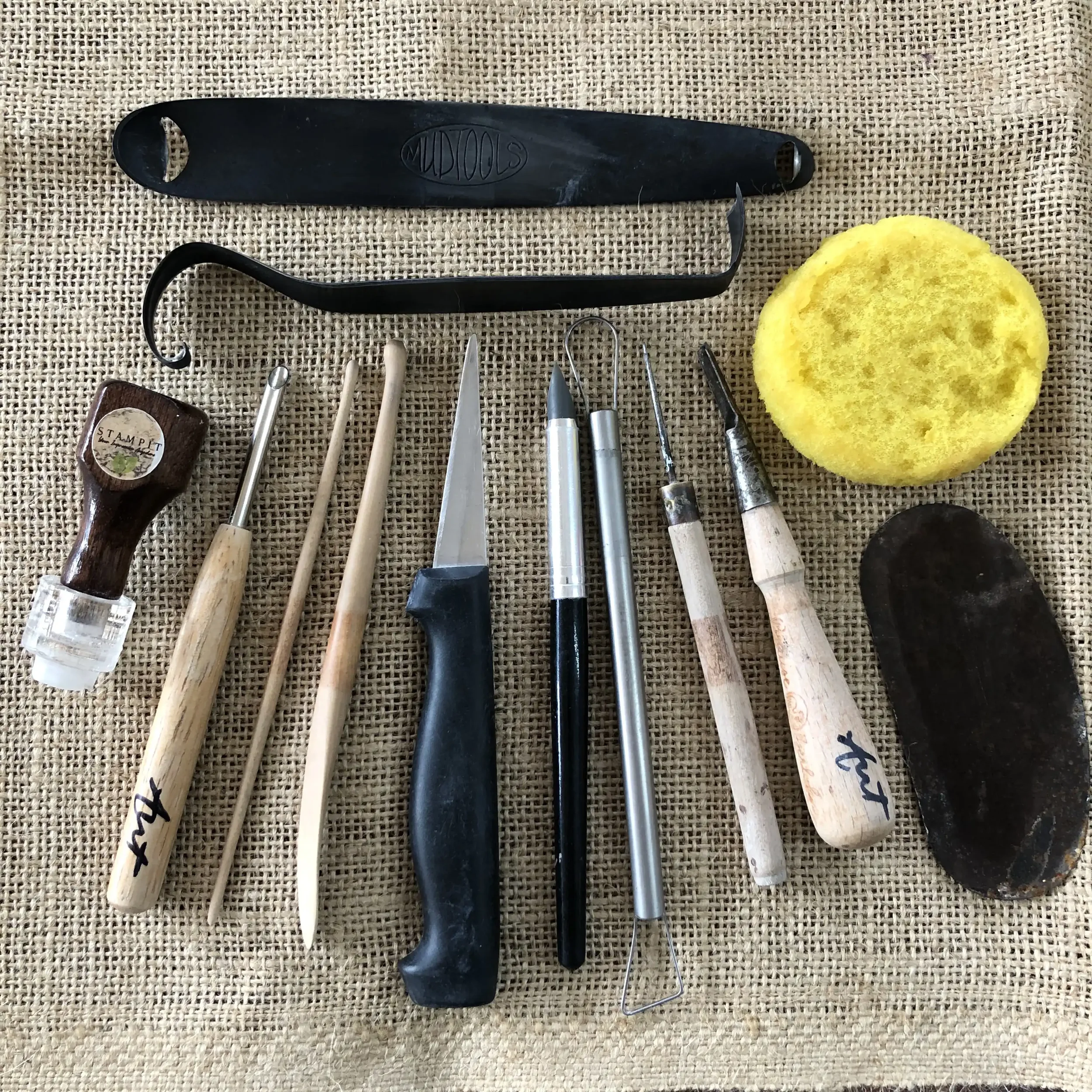 A variety of pottery tools arranged on a hessian surface, including knives, a sponge, and other wooden tools for pottery.