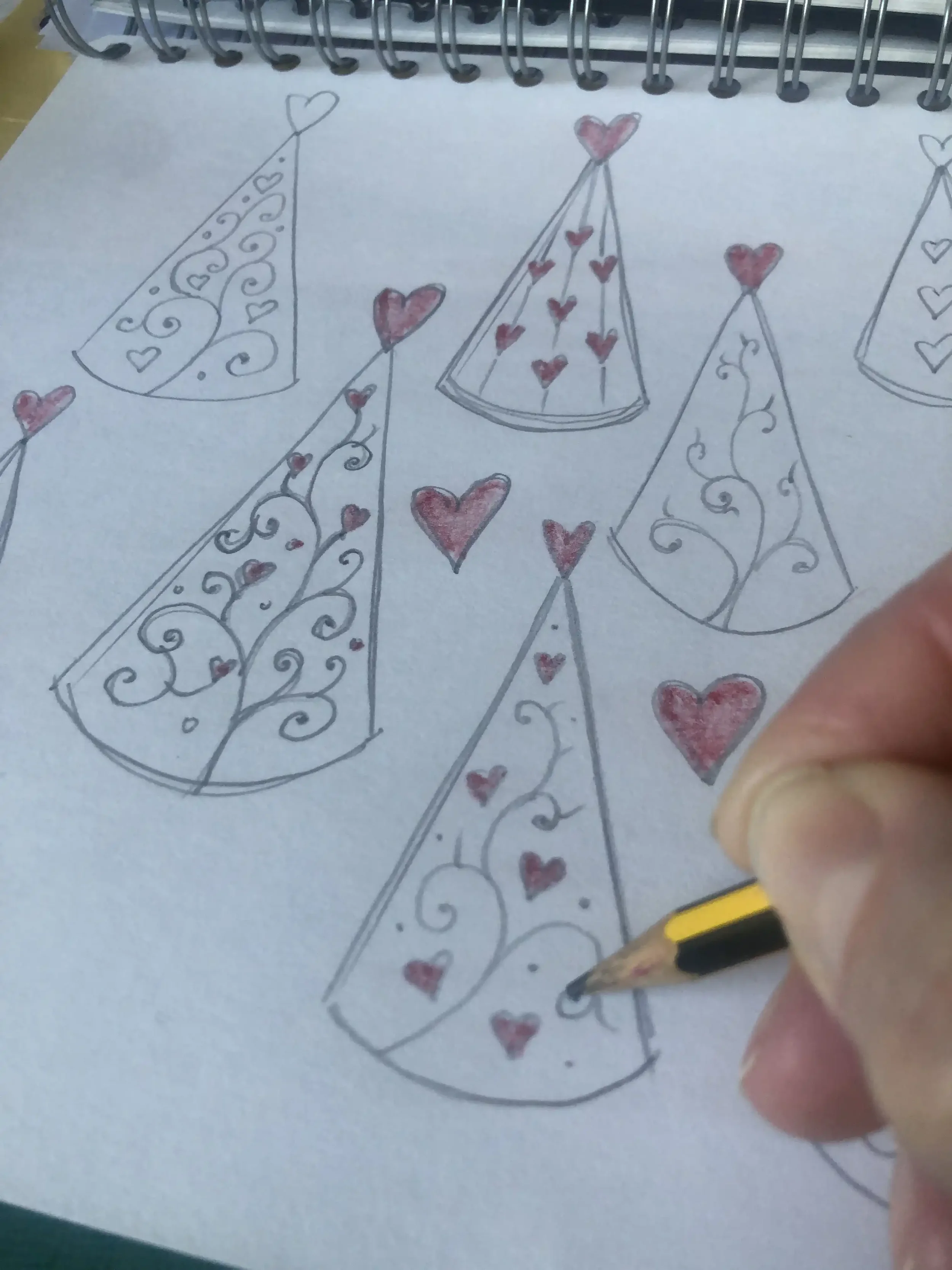 A person is drawing decorative cone shaped 'heart trees' with hearts and swirls on a sheet of paper with a pencil.