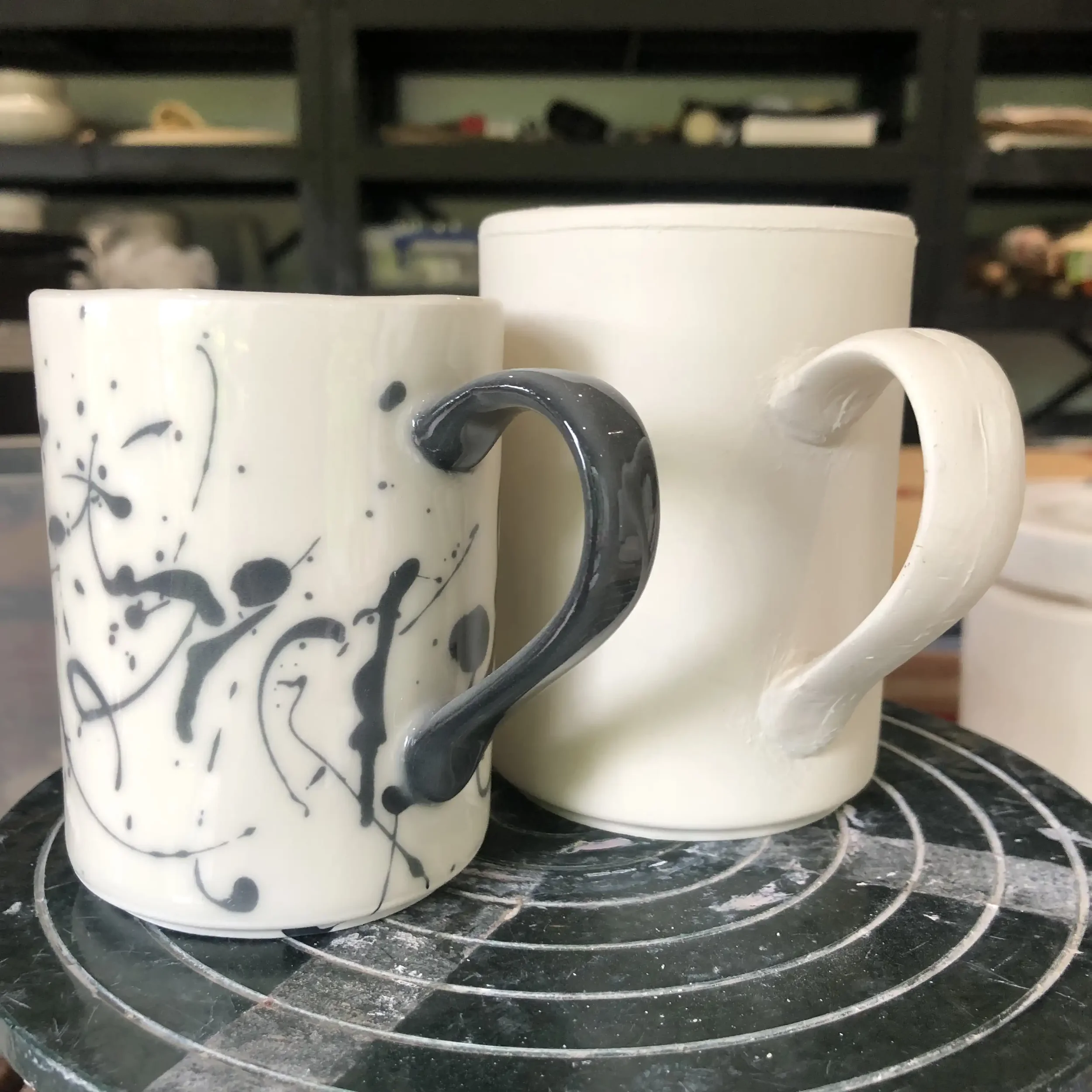 Two ceramic mugs sitting on a banding wheel, one with black and white abstract splashes and the other plain white.