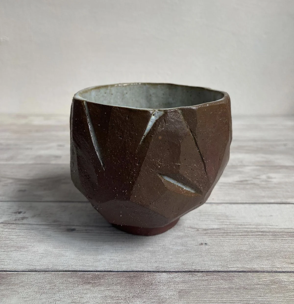 A geometric, handmade ceramic cup with a brown exterior and a light gray interior, placed on a light wooden surface.