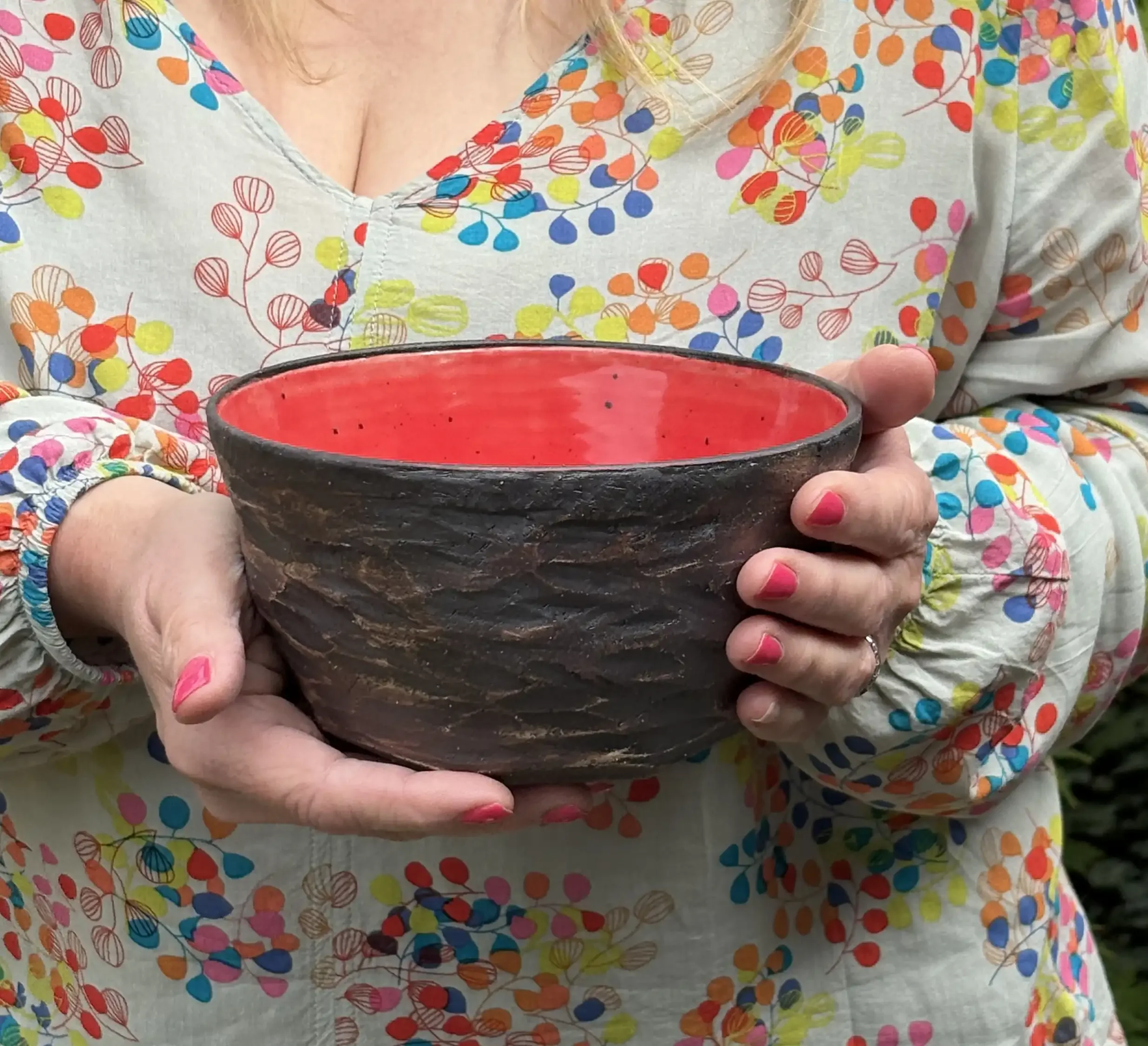 A woman wearing a colourful patterned blouse holds a ceramic bowl with a red interior and a textured, dark exterior.