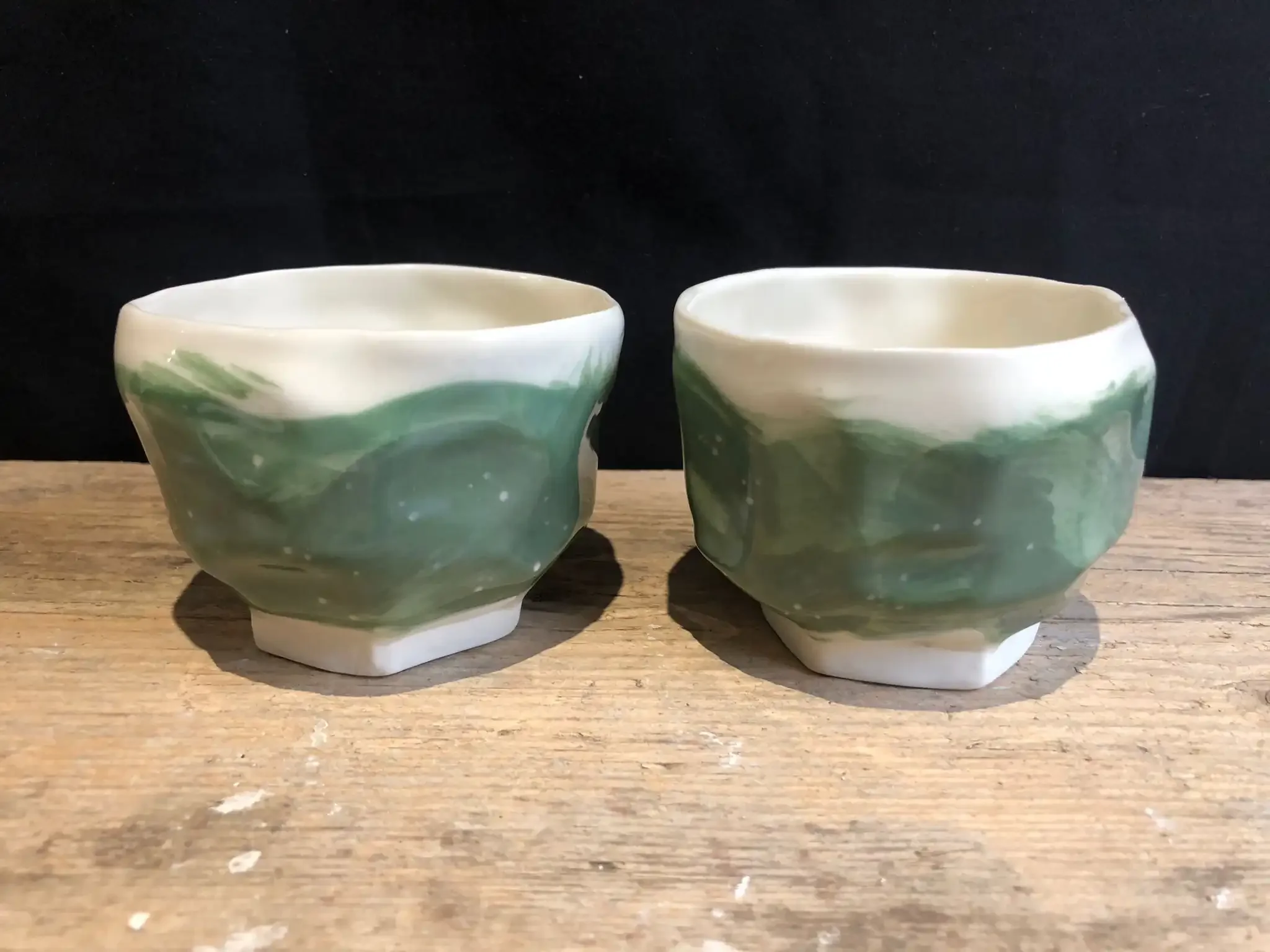 Two ceramic cups with green and white brushstroke designs with small white flecks sitting on a wooden surface against a black background.