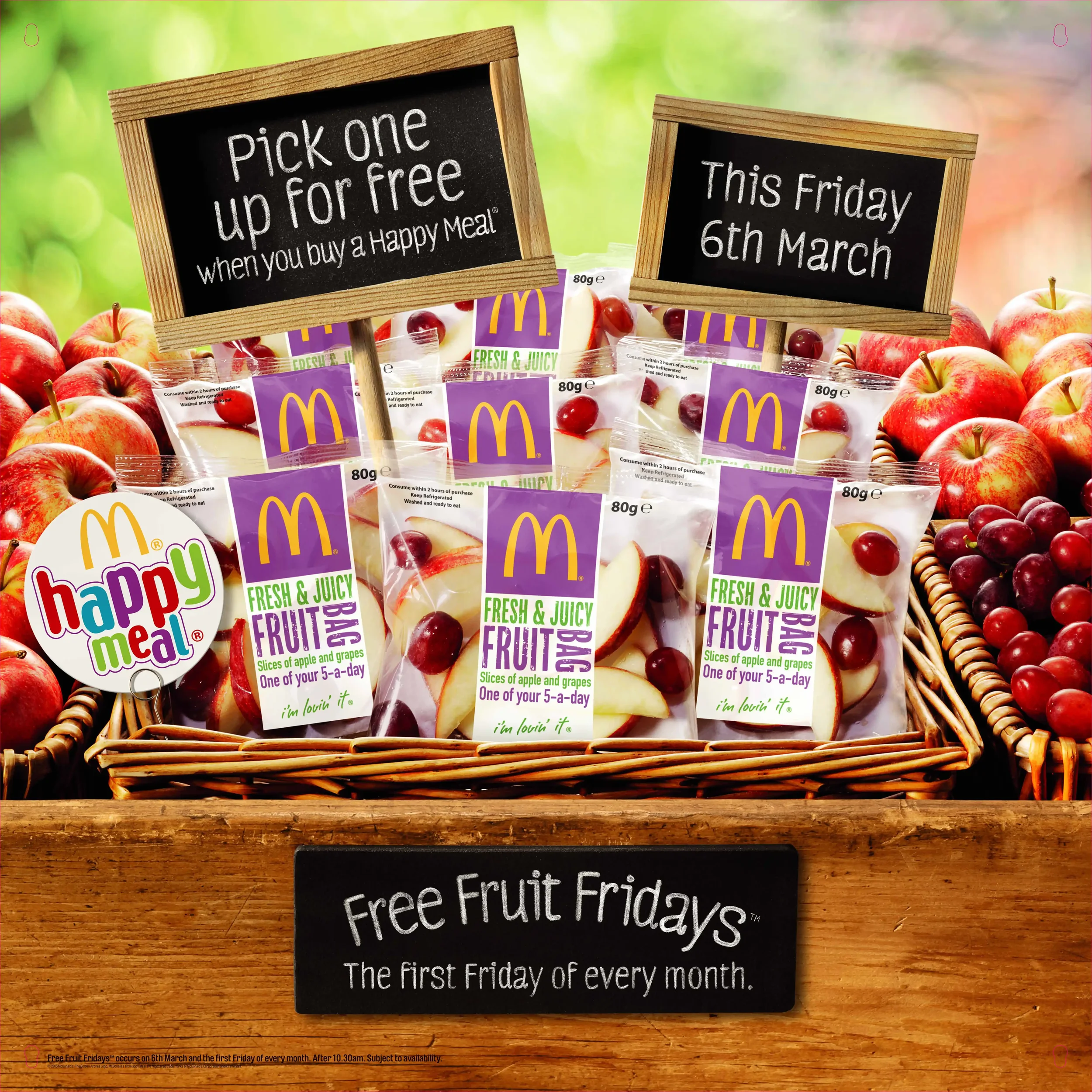 A display of McDonald's fruit snack packs labeled 'Fresh & Juicy Fruit,' showing slices of apple and grapes, with signs indicating a special offer for 'Free Fruit Fridays' on March 6th, placed among apples and grapes in a wooden basket.