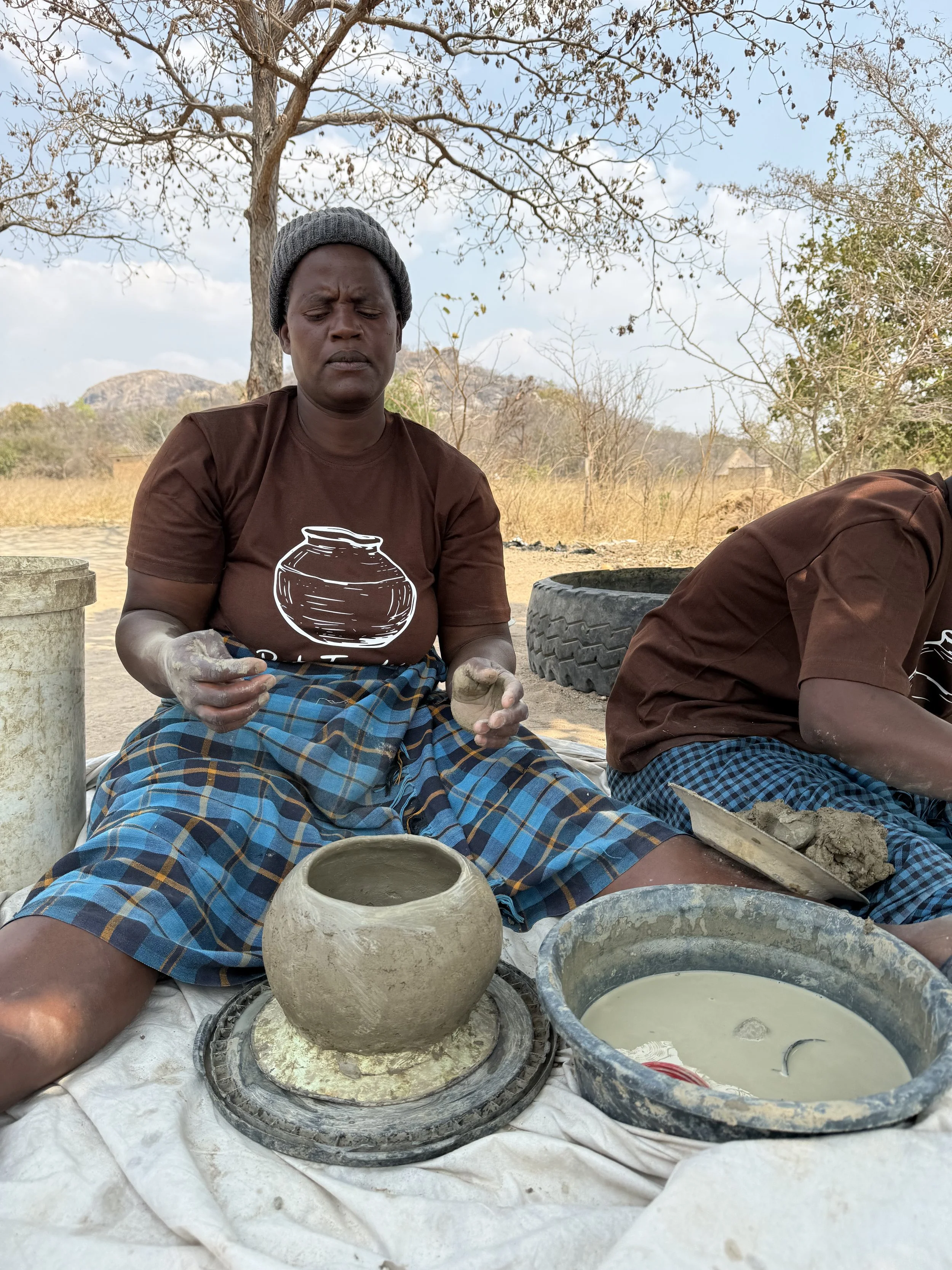A woman in a brown t-shirt with a pot graphic, plaid skirt, and gray beanie shapes clay with her hands while sitting outdoors on a white sheet, with bowls of clay and water, a tire, and trees in the background.