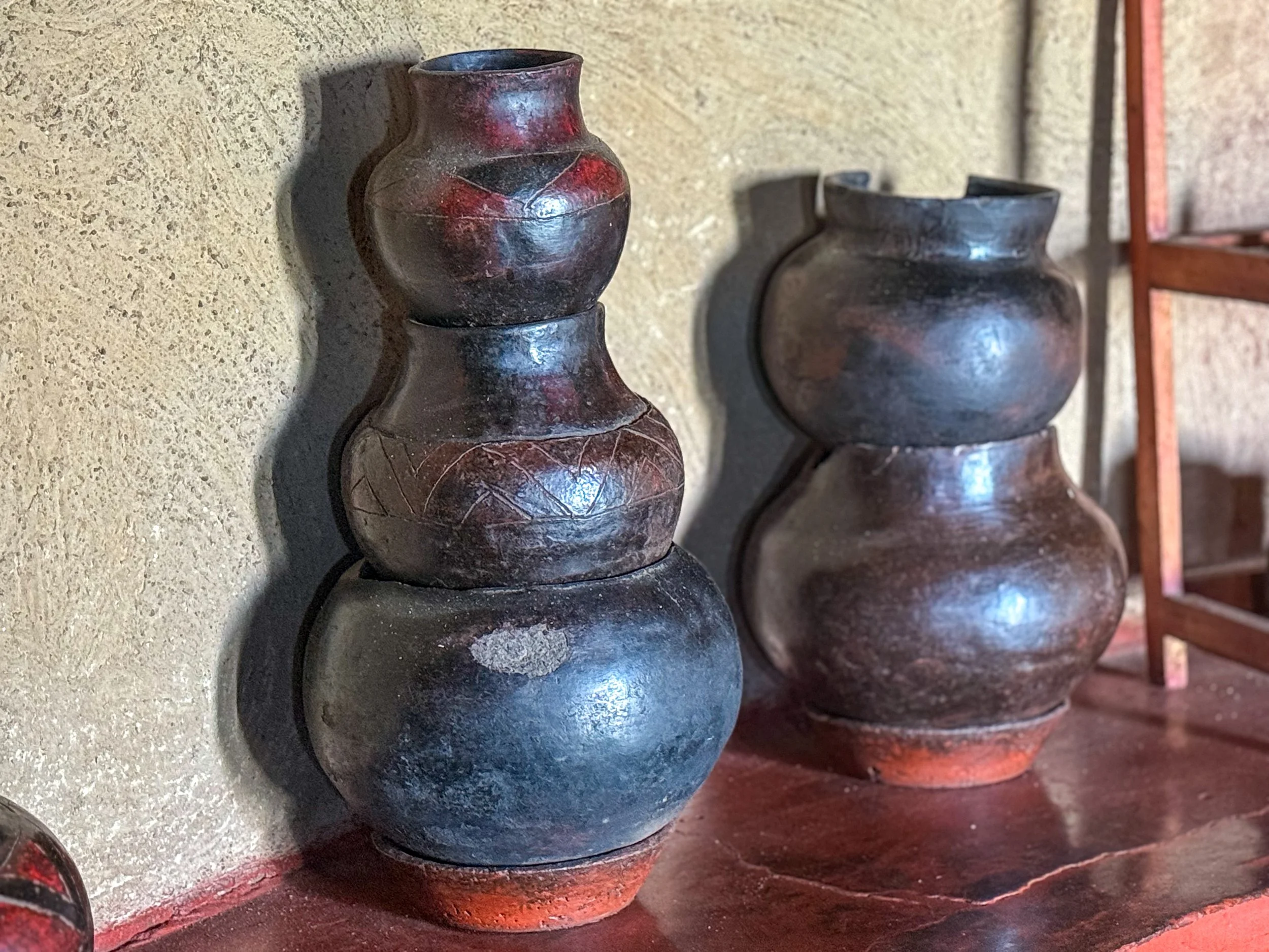 Two dark, rounded, stacked clay vases with a rough texture, placed on a wooden surface against a textured wall.