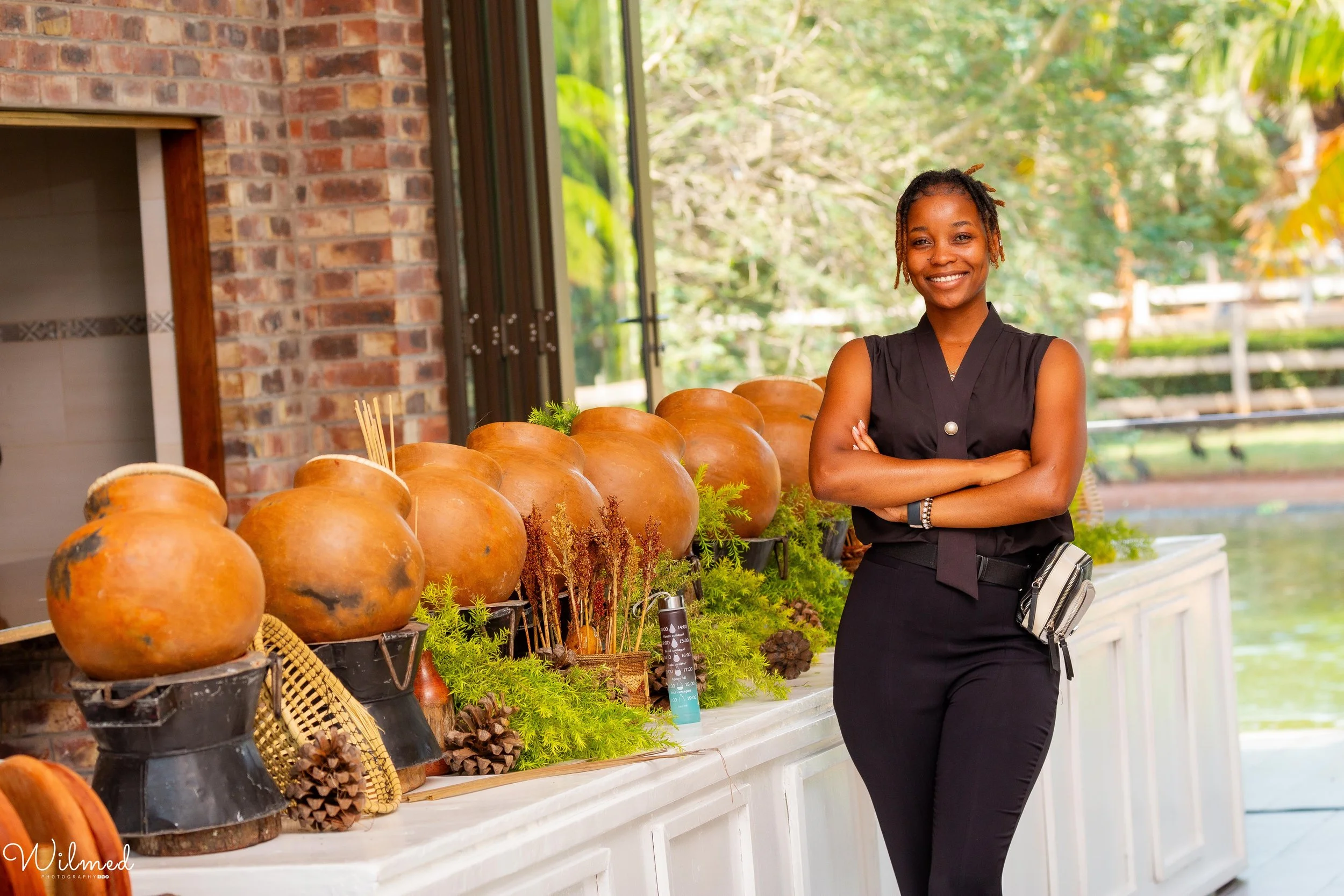 A smiling woman stands indoors near a display of large clay pots decorated with greenery and pinecones, with a brick wall and windows showing trees in the background.