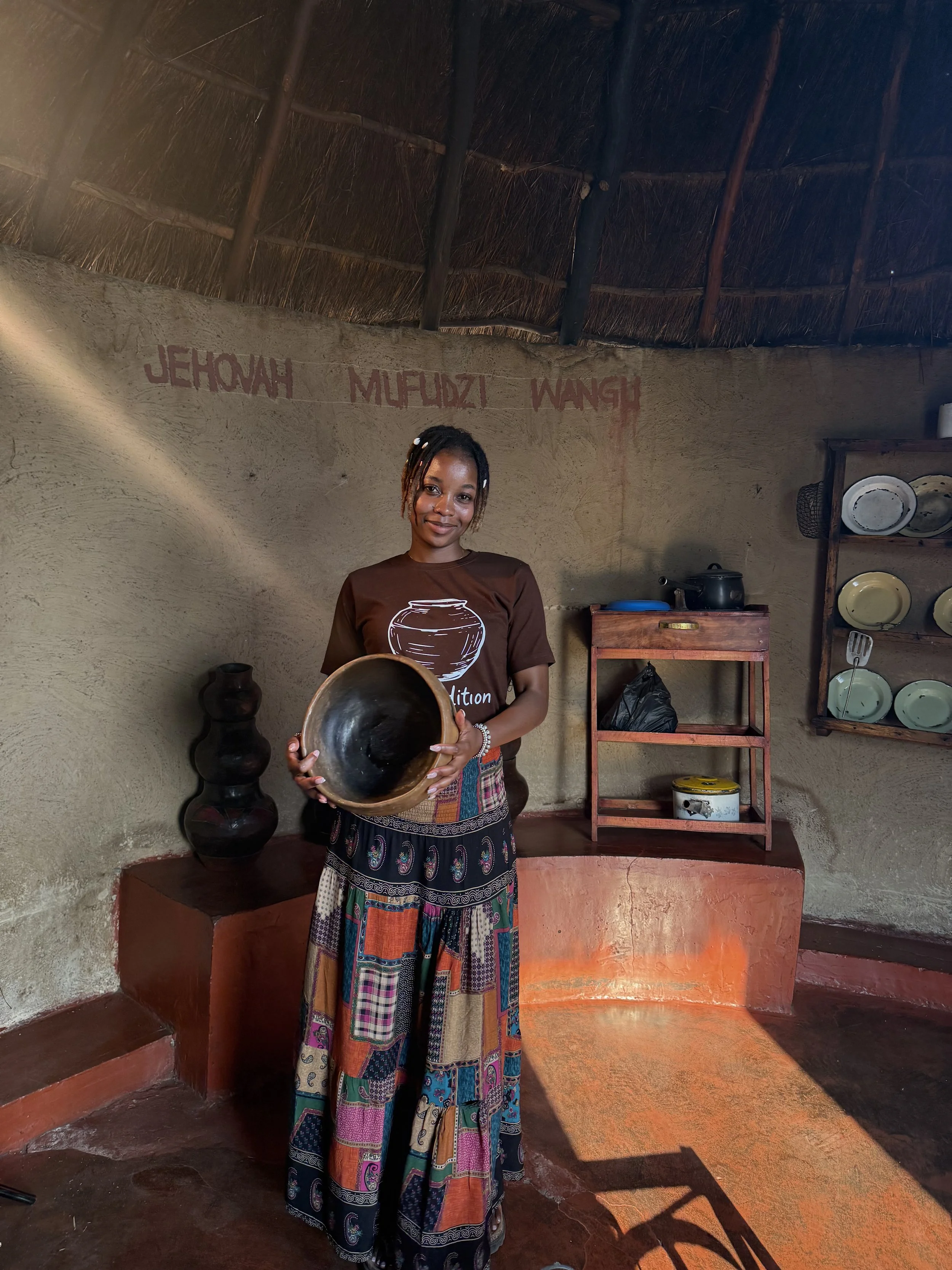 A woman standing inside a traditional hut, holding a black pottery bowl. The hut has a thatched roof and mud walls with red painted inscriptions. Shelves with pottery dishes and a small wooden table are visible in the background.