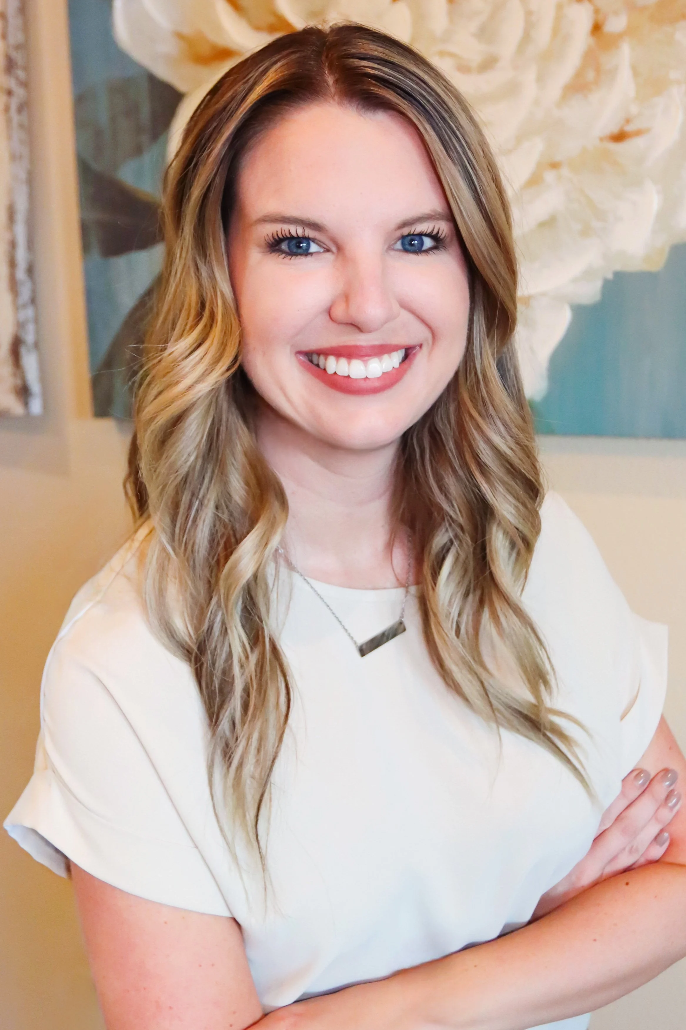 A woman with long wavy blonde hair and blue eyes smiling with arms crossed, wearing a white top and a silver necklace in front of a large floral artwork.