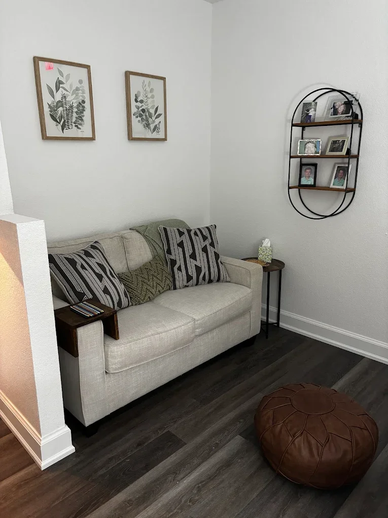 A cozy living room corner with a beige couch adorned with patterned cushions, a small wooden side table with a tissue box and remote, a wall shelf holding framed photos, and a brown leather pouf on dark wood flooring.
