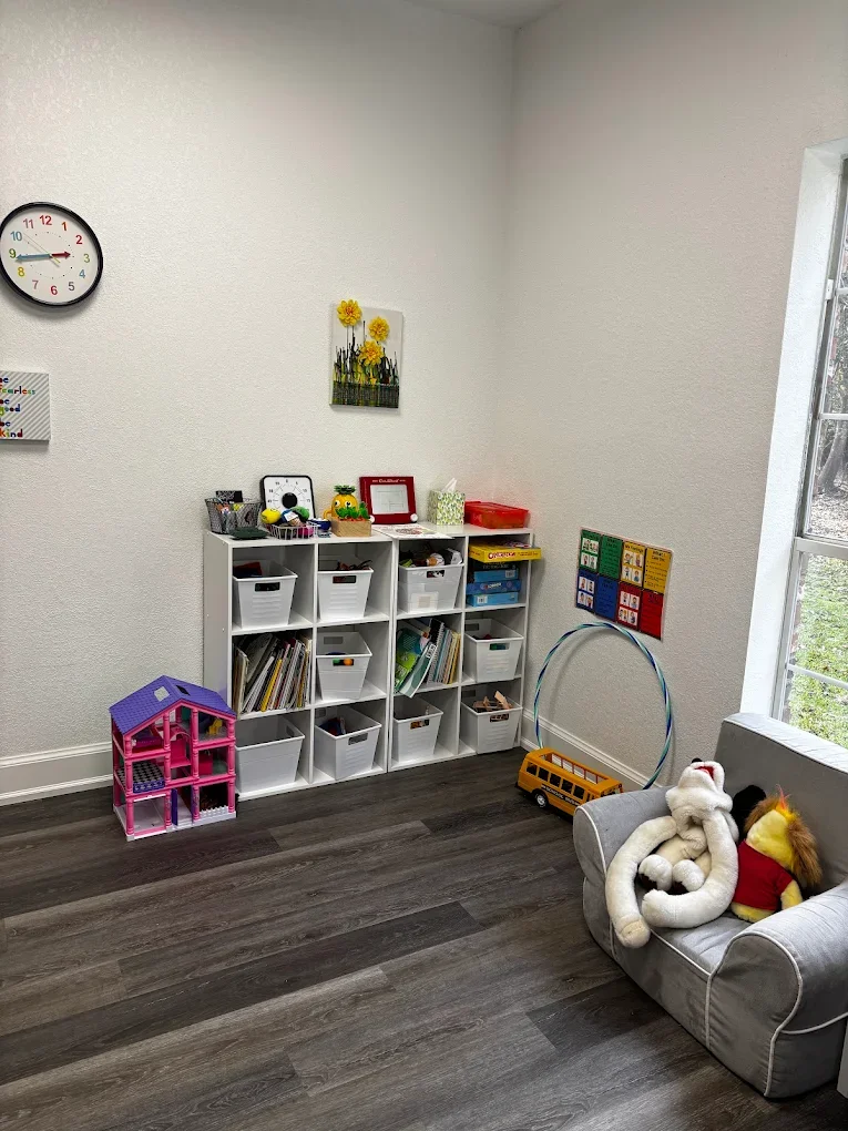 A corner of a children's playroom with a white cubical storage unit holding books and toys, a pink dollhouse, a yellow toy school bus, and plush toys on a small gray armchair near a window, with wall art and a clock on the wall.