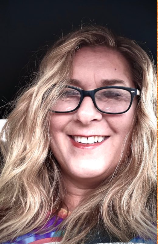 A woman with long blonde wavy hair and black glasses smiling at the camera.