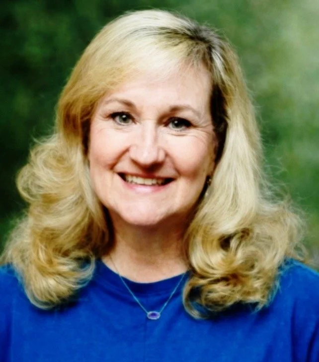 A middle-aged woman with blonde hair and a warm smile, wearing a blue top and a necklace, against a blurred green background.