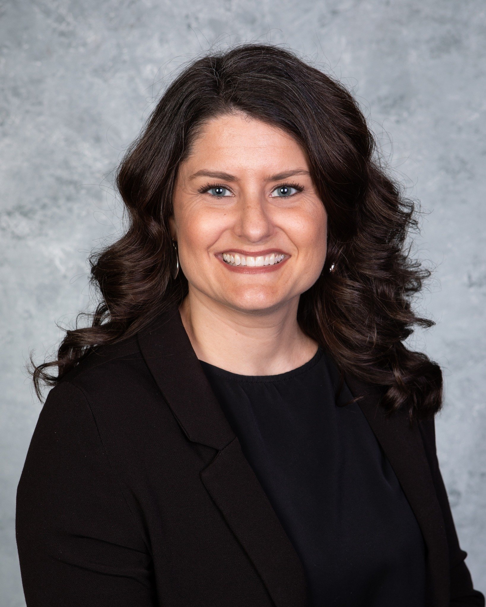 Professional headshot of a woman with shoulder-length dark wavy hair, wearing a black blazer and black top, smiling in front of a gray textured background.