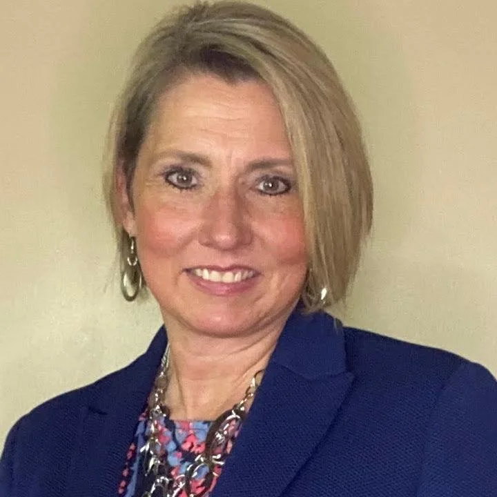 A woman with short blonde hair wearing a dark blue blazer, colorful necklace, and earrings, smiling at the camera against a plain beige background.