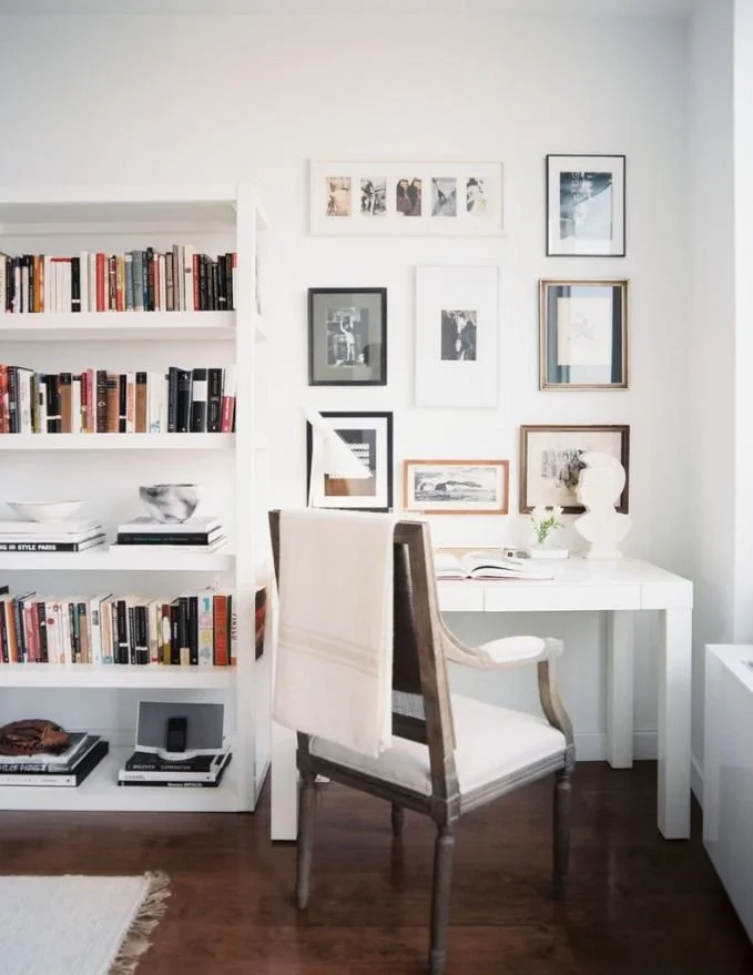 A cozy, well-lit home office with a white bookshelf filled with books on the left and framed art on the wall above a white desk on the right. A cushioned chair faces the desk, which has an open book and decorative sculptures, with a dark wooden floor.