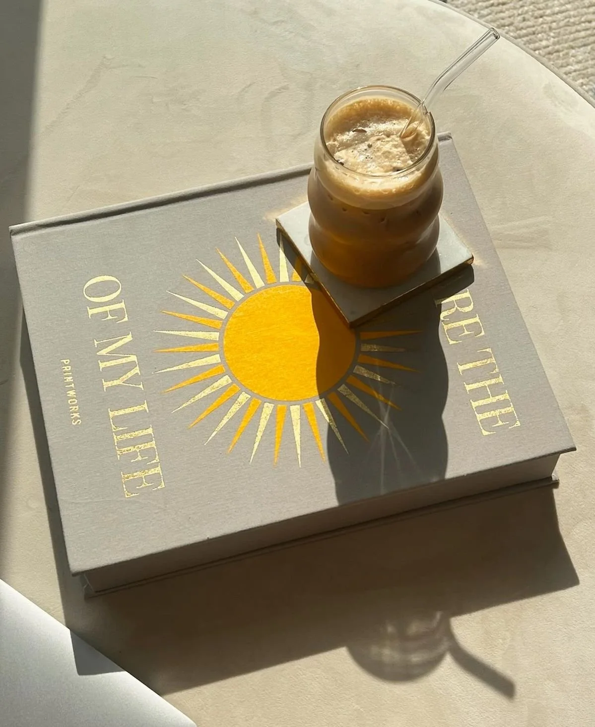 A glass of iced coffee with a straw placed on a book titled 'Reclaim Your Time' by Philtworks, sitting on a white surface with sunlight casting a shadow.