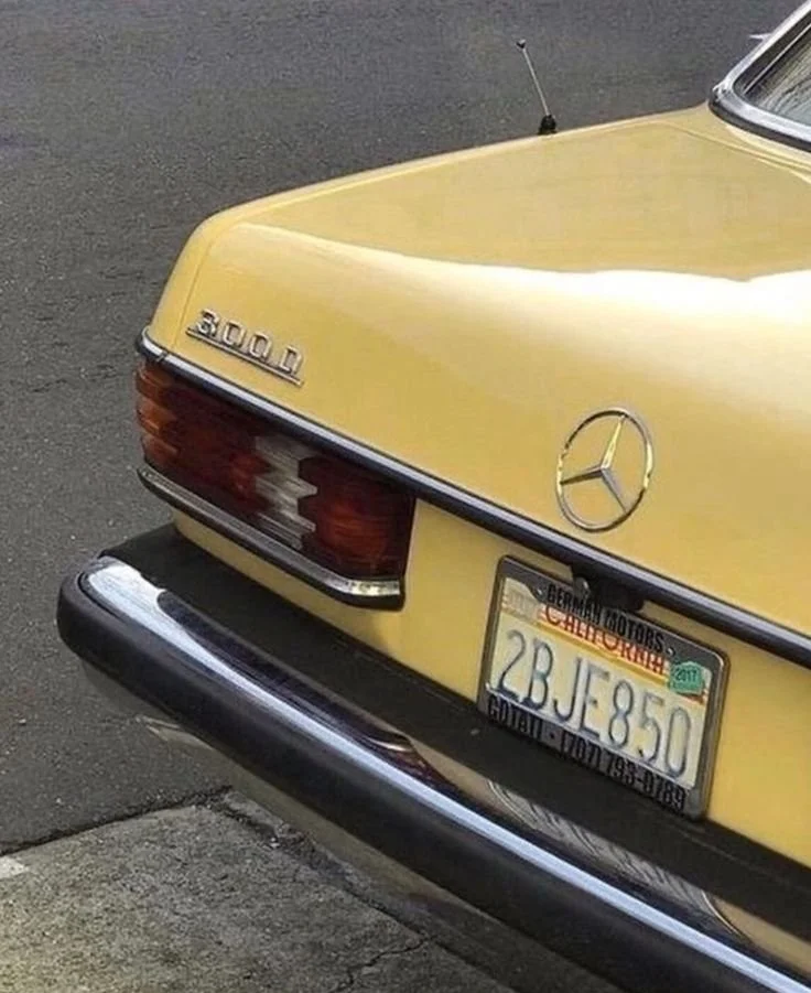 The rear of a yellow Mercedes-Benz 300D car parked on the side of the road, showing the car's badge and license plate.