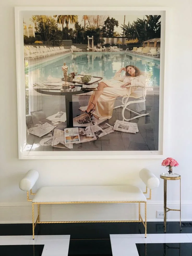 Decorative framed photograph of a woman relaxing by a swimming pool, surrounded by chairs and lush greenery, mounted on a wall above a white and gold bench with a small table holding pink roses.