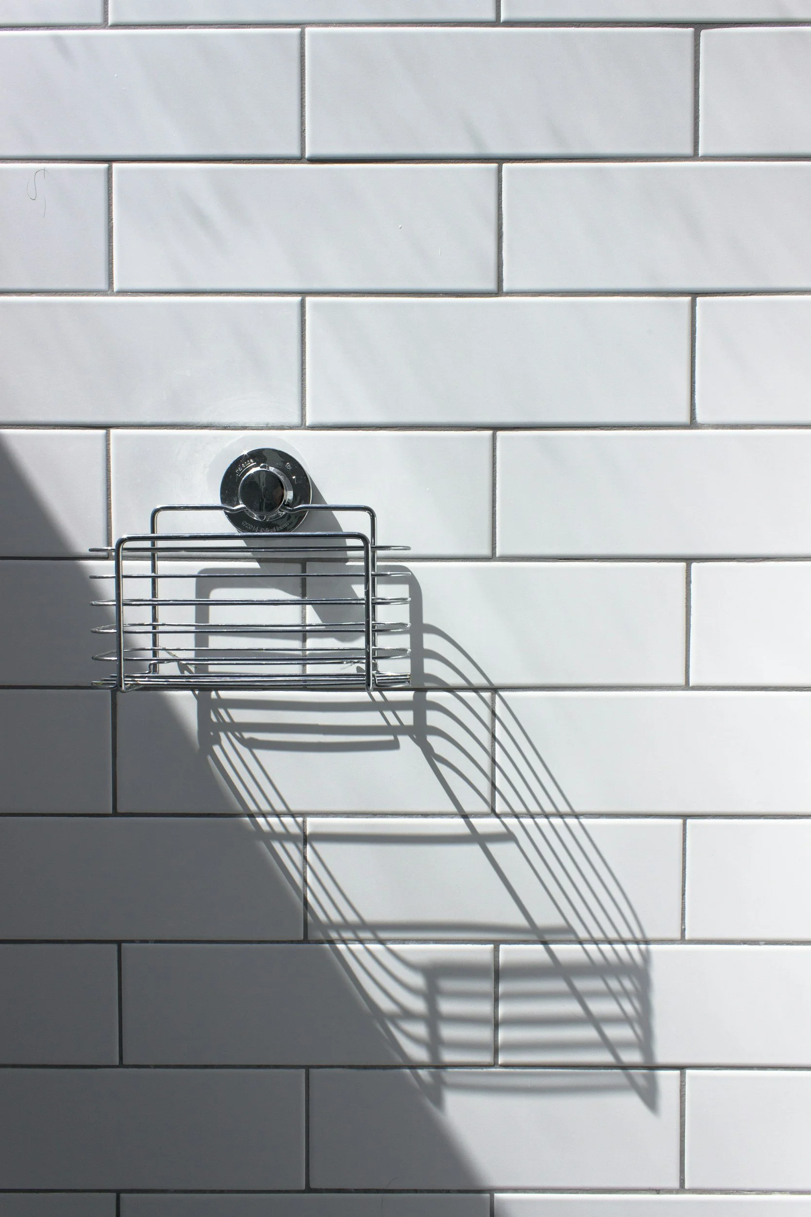 A shower wall with white subway tiles, a metal wire soap holder, and sunlight casting a shadow of the soap holder on the tiles.