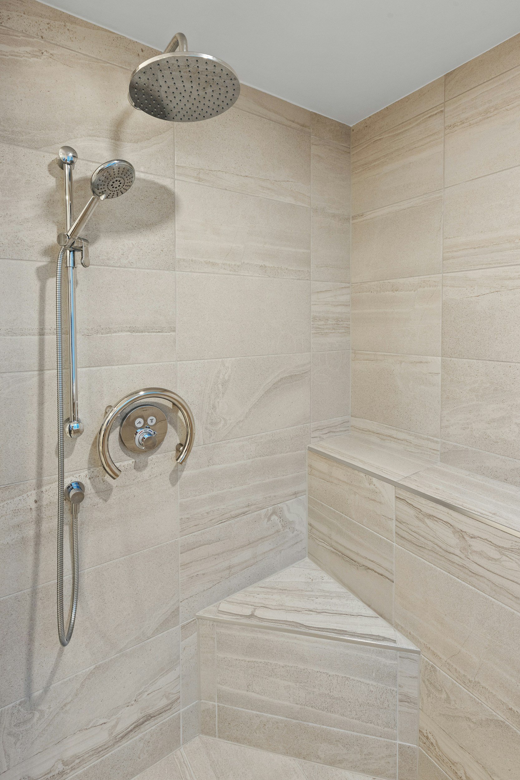 Shower area with two shower heads and beige tiled walls and bench.