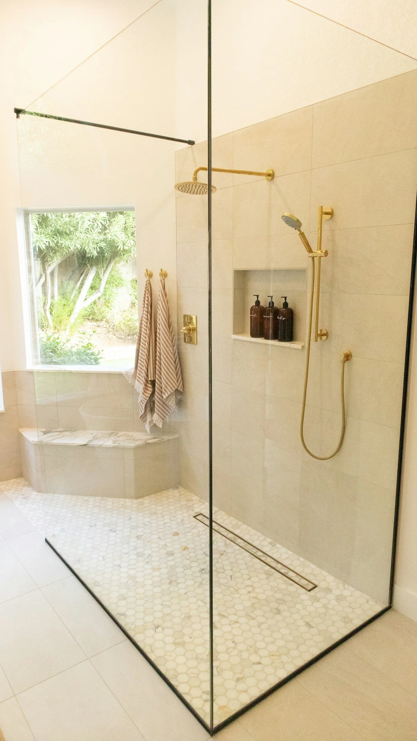 Modern walk-in shower with beige tiled walls and gold fixtures, including a shower head, handheld spray, and a built-in niche with brown bottles. The shower floor has small white hexagonal tiles and a linear drain. Adjacent to the shower is a window 