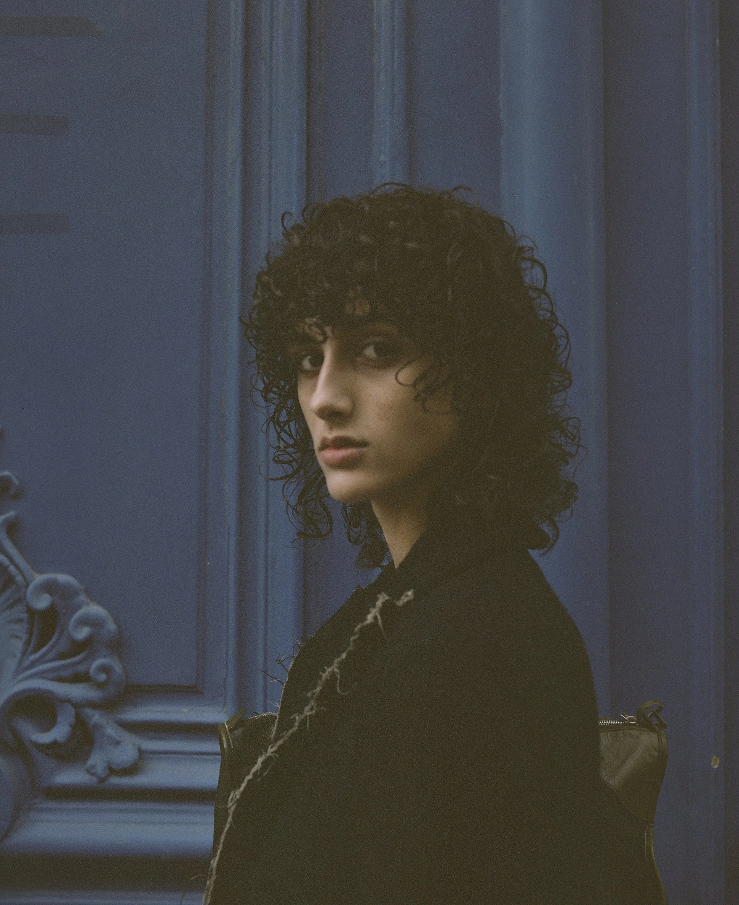 A young person with dark, curly hair and a serious expression, wearing a black jacket, standing against a blue decorative wall with ornate details.