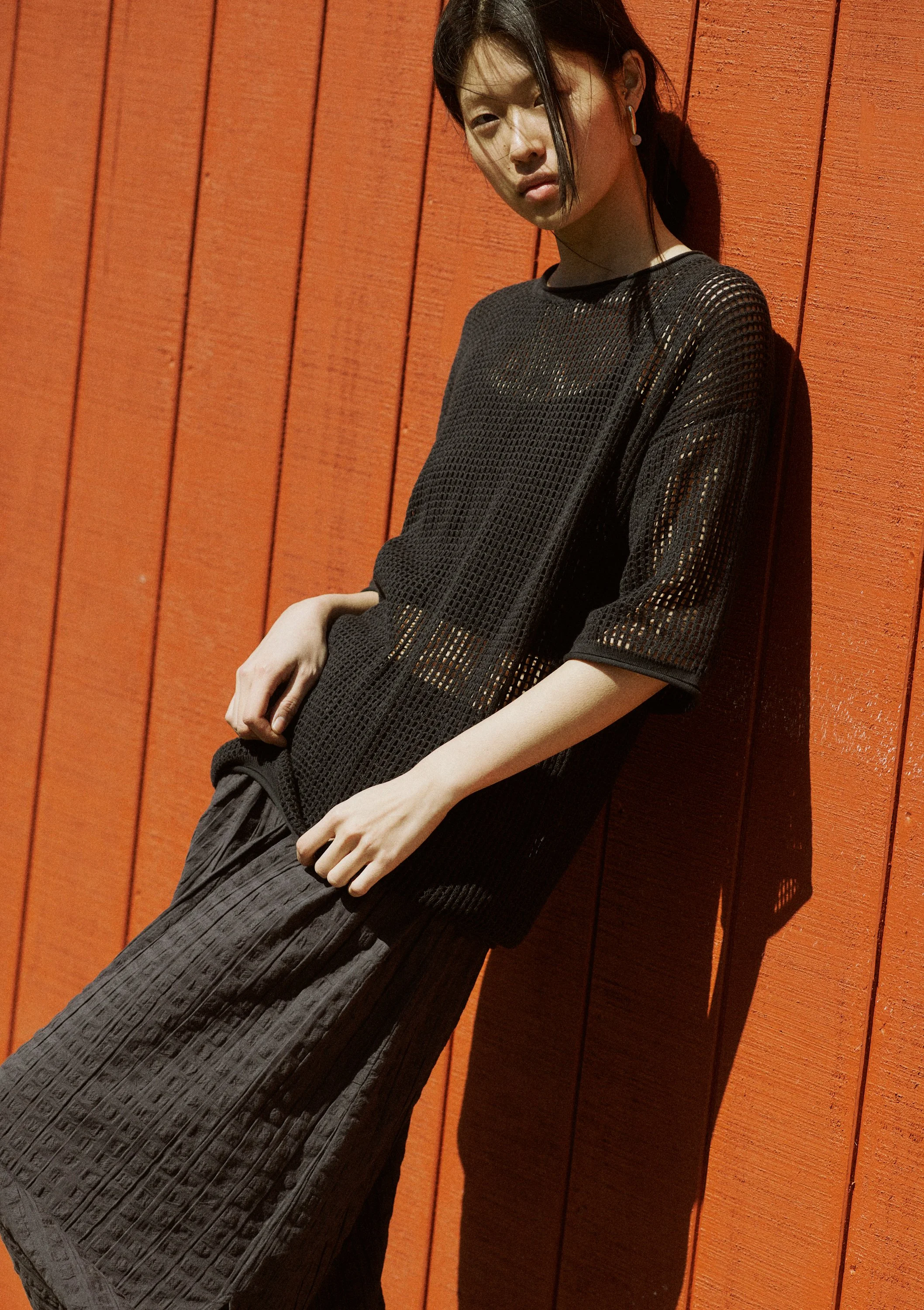 Young woman with dark hair leaning against a red wooden wall, wearing a black mesh top and a textured black skirt.