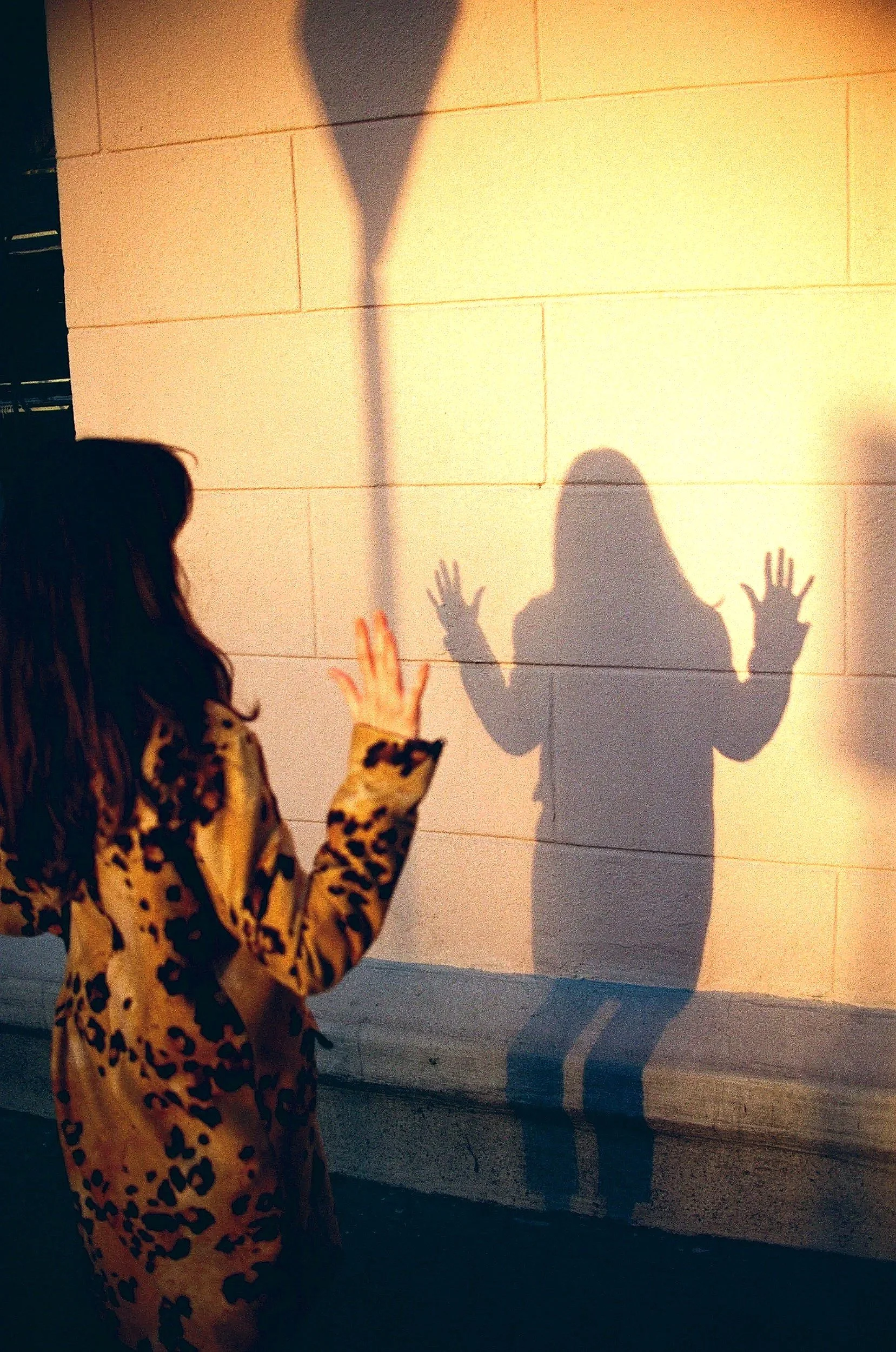A woman in a leopard print coat touches a wall with her hand as her shadow is cast onto the wall, creating an illusion of a person with raised hands.