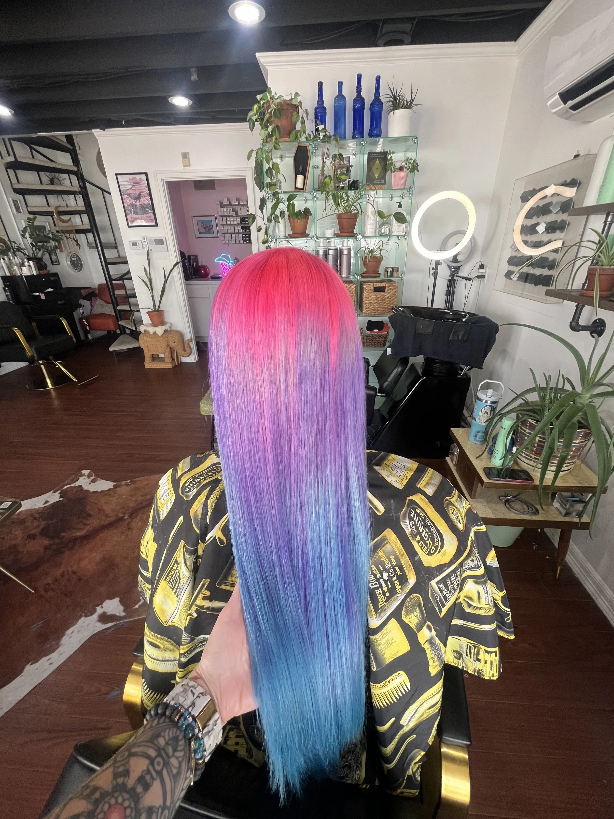 A person with long, straight hair dyed in a gradient of pink, purple, and blue, sitting in a salon chair with a black and yellow cape. The salon interior features plants, decorative bottles, and ring lights.