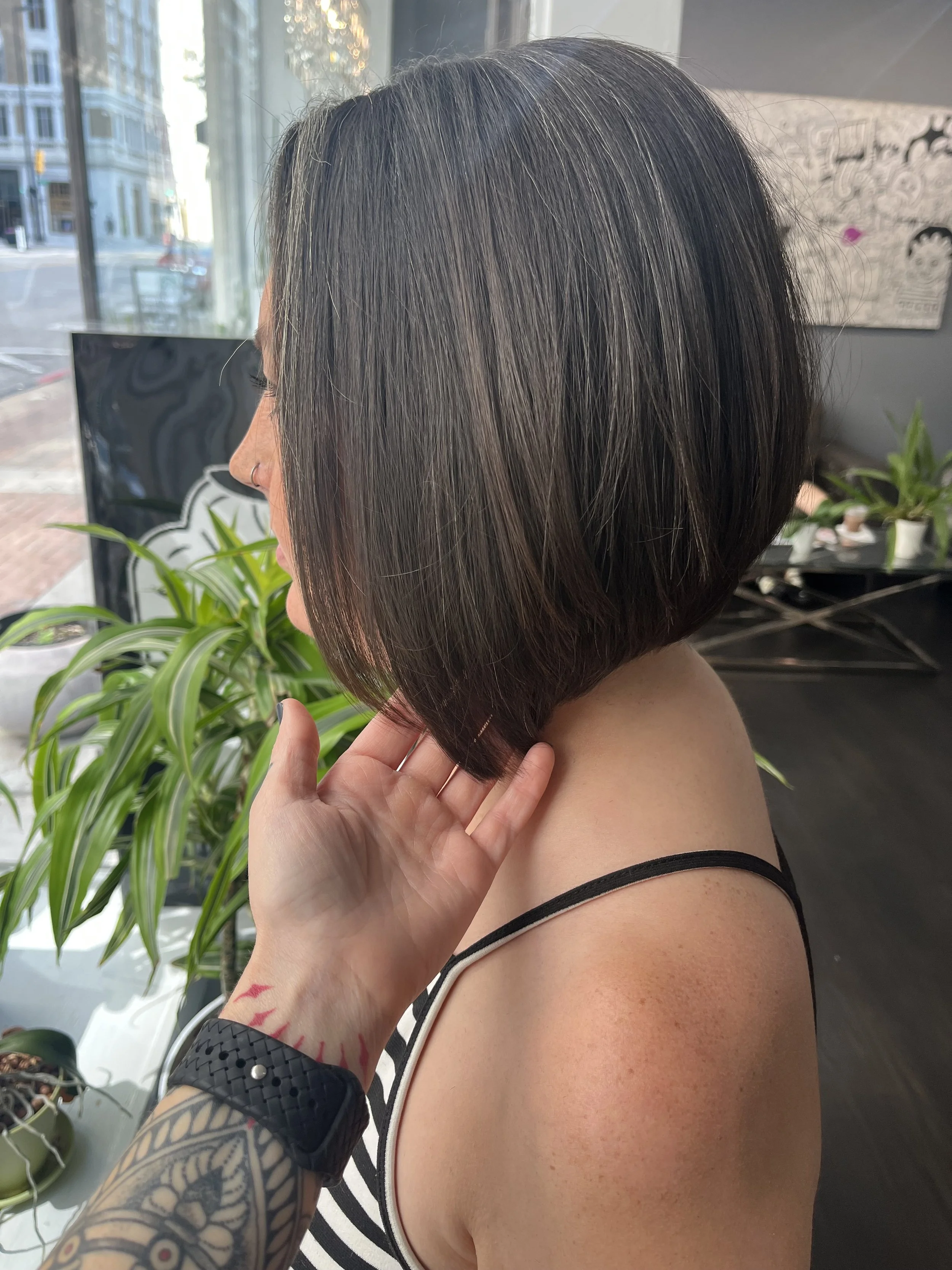 A woman with a short, dark bob haircut sits inside a salon near a large window, with a tattooed arm touching her neck, wearing a black and white striped top.