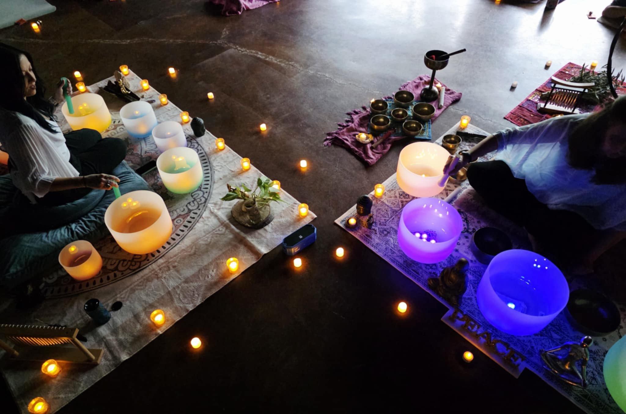 Two people seated on colorful mats, playing singing bowls under dim lighting with candles and decorative items around them.