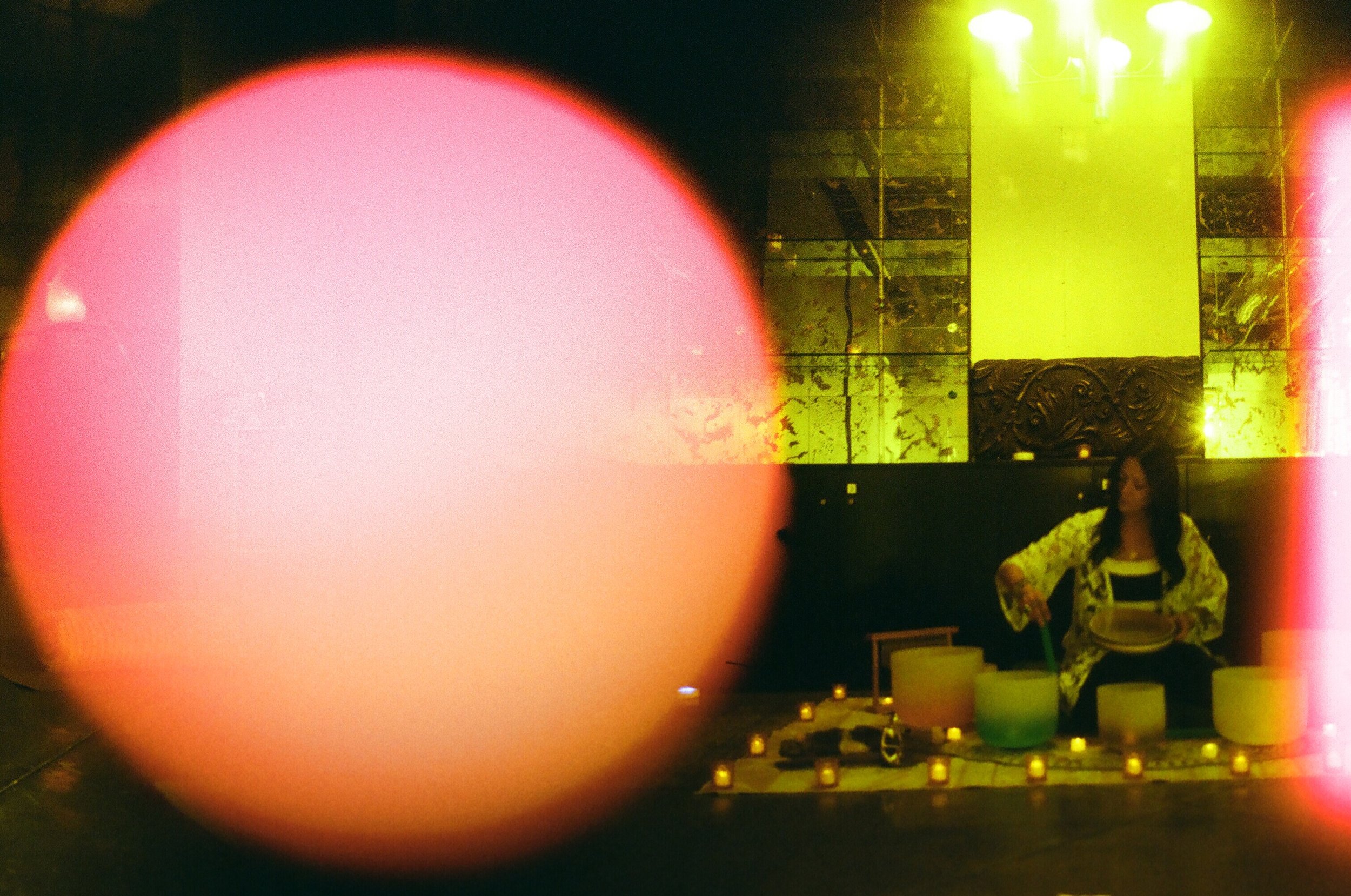 A woman sitting in a dimly lit room with yellow lighting, surrounded by candles and musical instruments, with a large pinkish light reflection or lens flare in the foreground.