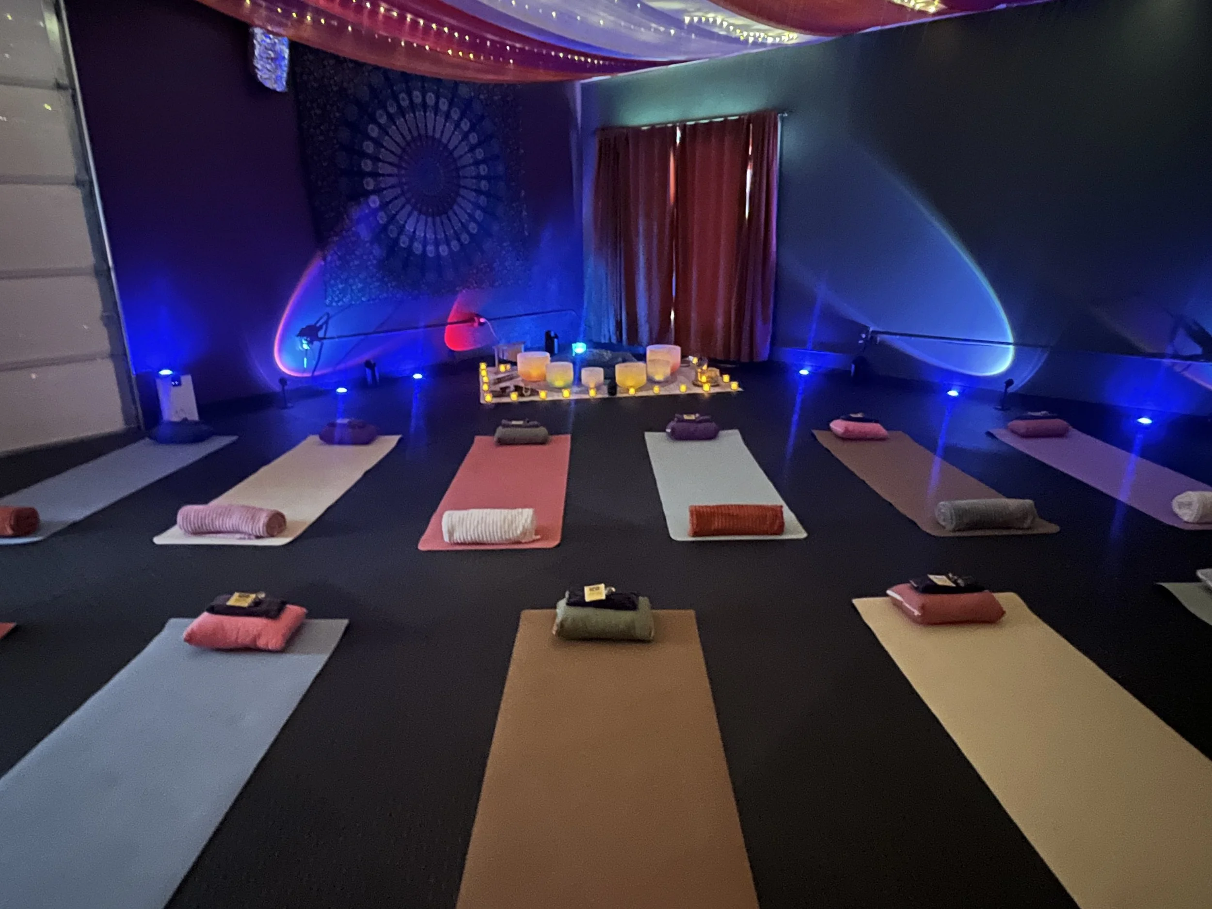 Yoga mats and towels arranged in a circle around candles in a dimly lit room for a meditation or yoga class, with colorful lighting and decorative wall art.
