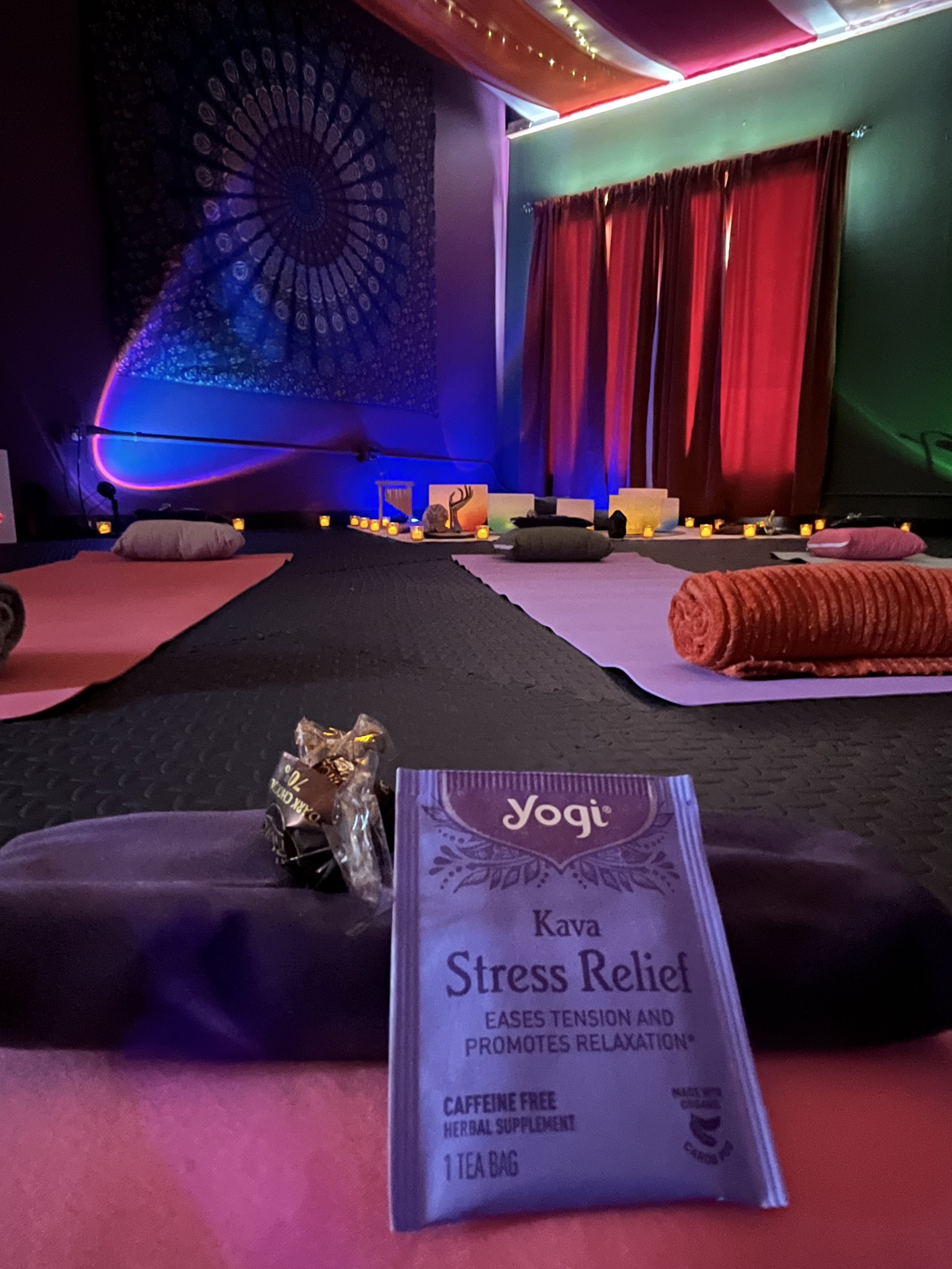 A yoga studio with candles, bolsters, and peaceful lighting, featuring a colorful tapestry on a wall and red curtains.