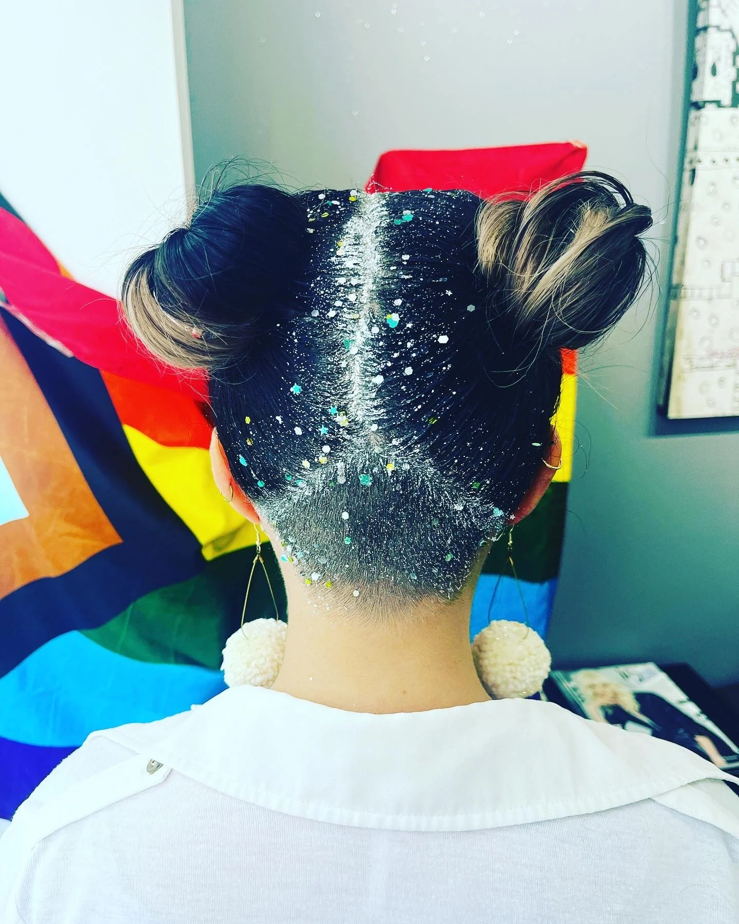 Back of a person's head with a hairstyle featuring two buns and a shaved undercut, decorated with white and multicolored paint splatters, against a rainbow pride flag background.