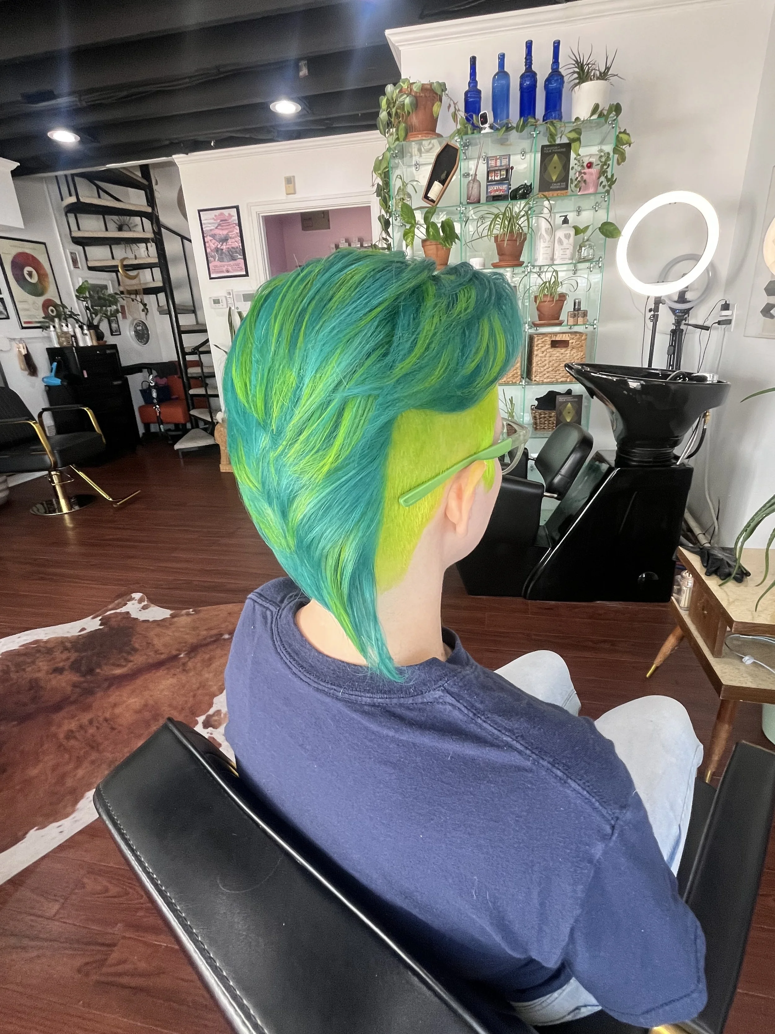 Person with vibrant green and blue hair and green glasses sitting in a salon chair.