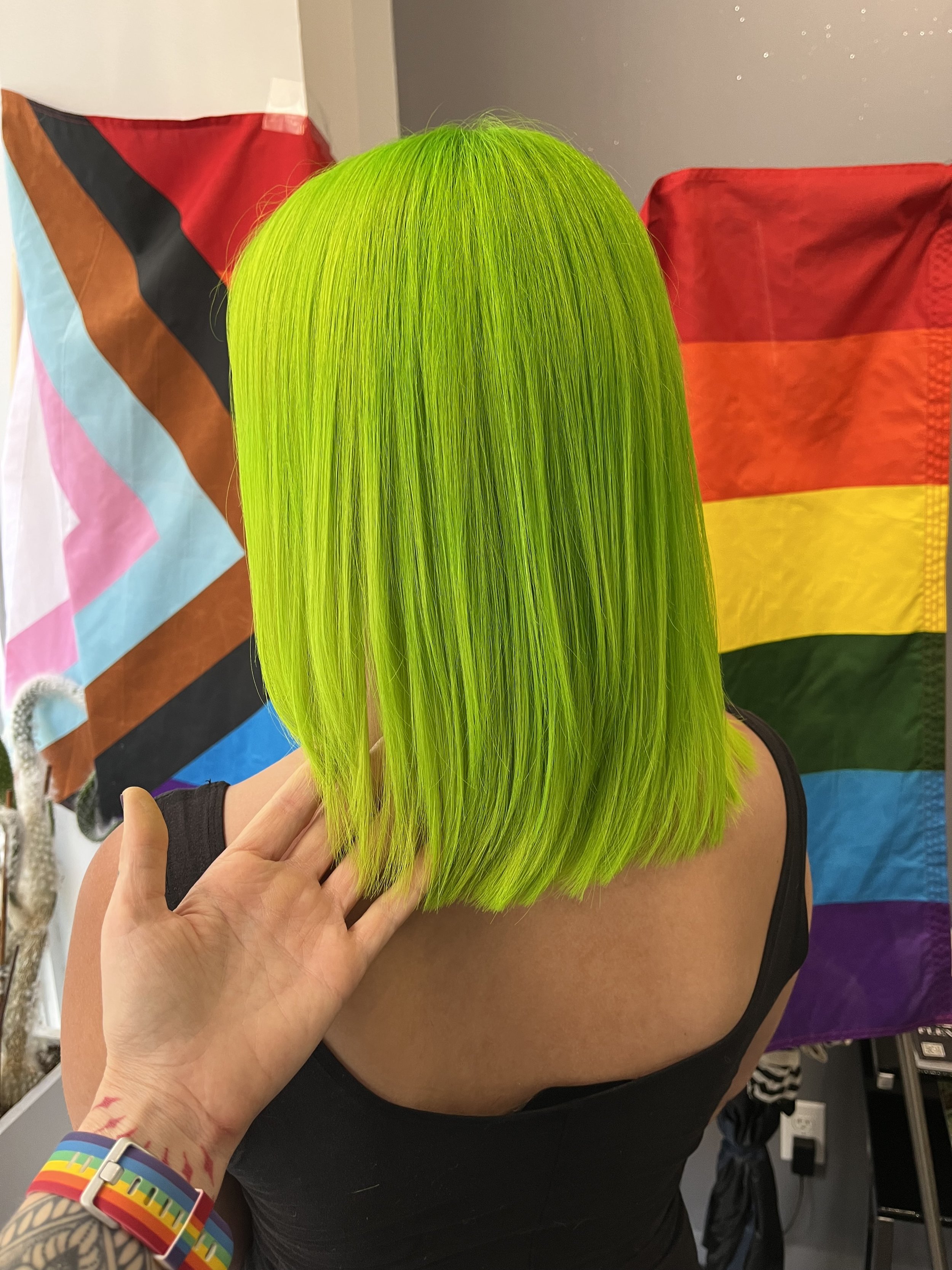 Person with vibrant neon green straight bob haircut posing with hand near hair, with rainbow flag and a pride flag in the background.