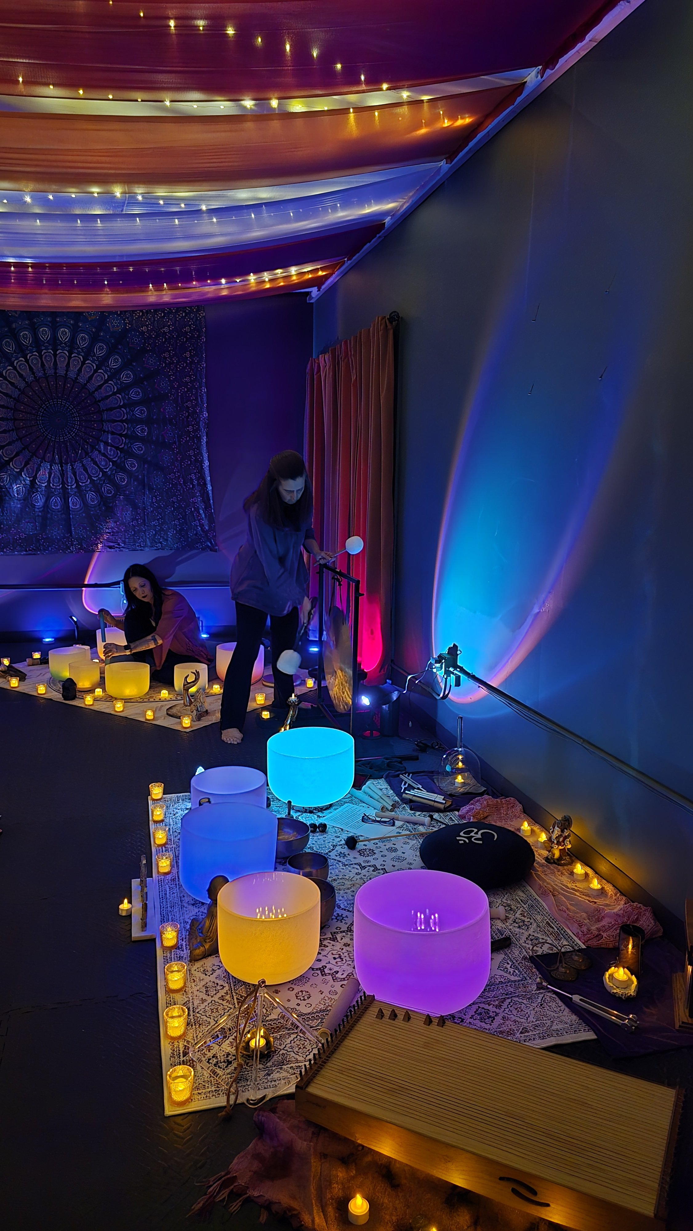 Two women in a colorful, dimly lit room with musical instruments and glowing lamps, preparing for a music or meditation session.