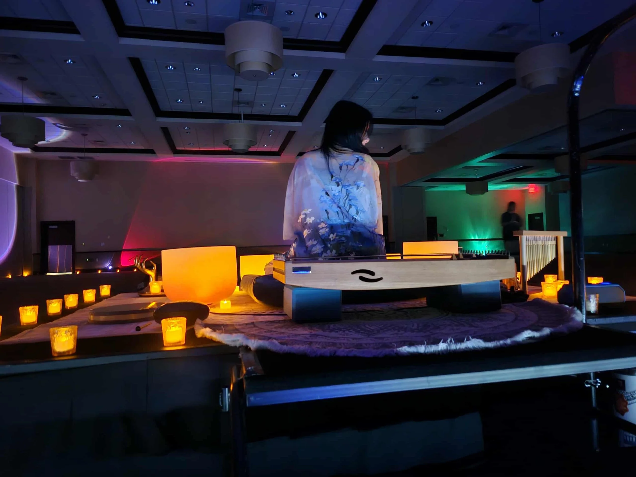 A woman with dark hair in a ponytail sits at a DJ mixing console on a stage decorated with candles, illuminated by colorful ambient lighting in a large indoor event space.