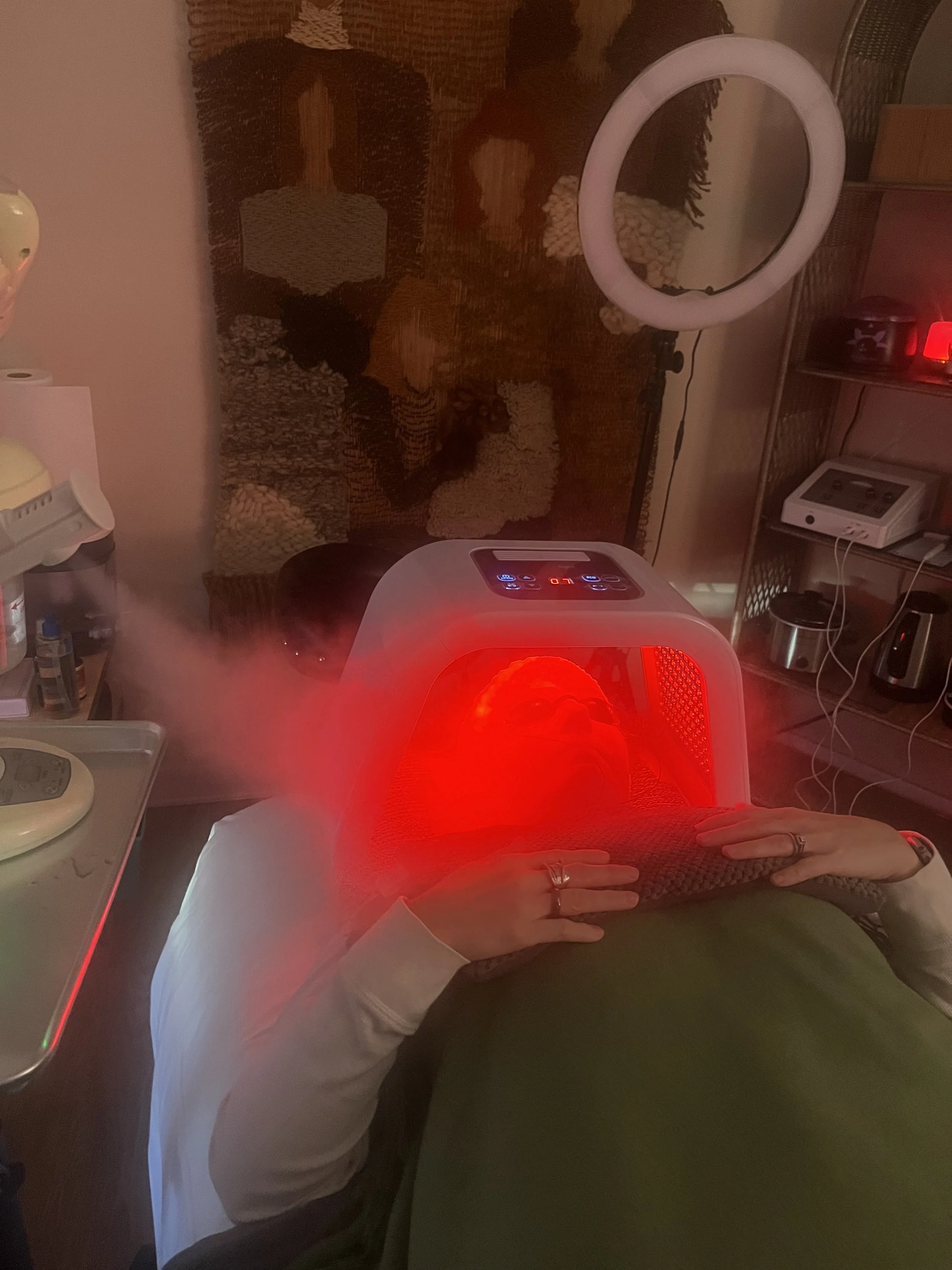 Person undergoing red light therapy from a light therapy device in a home setting, with a tapestry on the wall and various kitchen appliances visible.