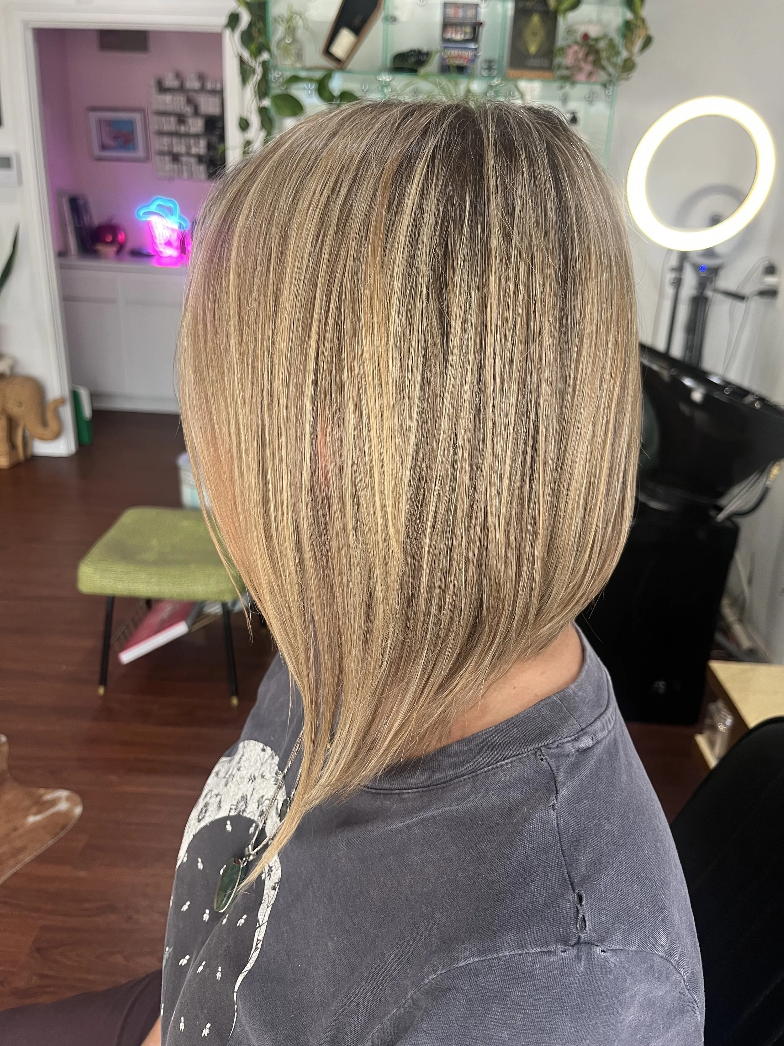 A person with shoulder-length blonde hair sitting in a hair salon with a ring light and salon equipment visible in the background.