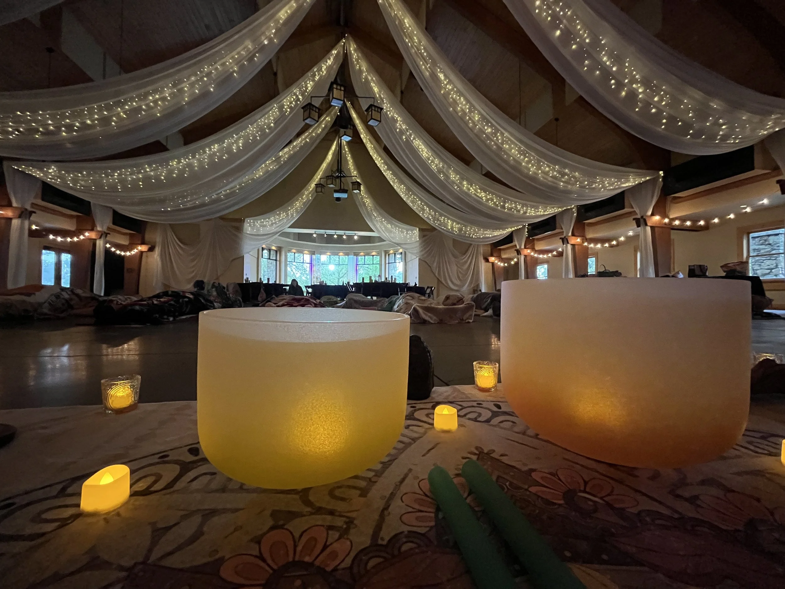 An interior decorated for a celebration with white draped fabric and string lights on the ceiling, large windows at the far end, and illuminated candles and singing bowls placed on a table in the foreground.