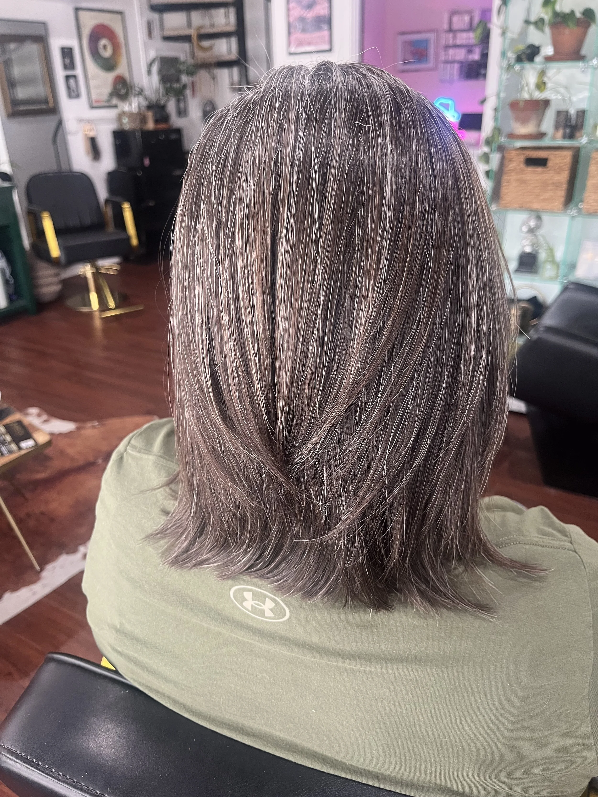 Back of a person's head with shoulder-length, layered hair with gray and dark brown highlights, sitting in a salon with shelves of decor and equipment in the background.