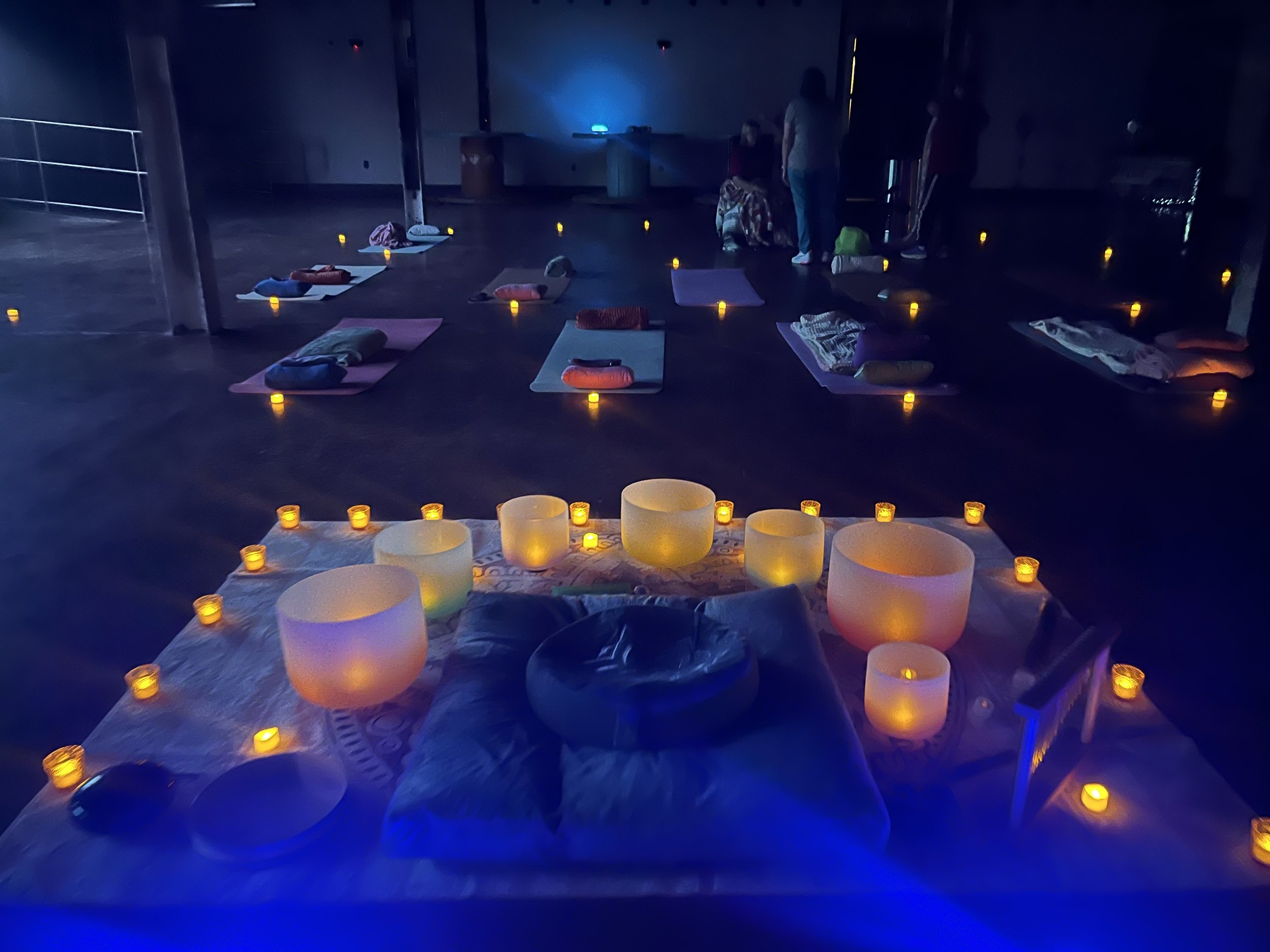 A dimly lit yoga or meditation studio with mats and blankets arranged on the floor, surrounded by small candles, and Tibetan singing bowls in the foreground.
