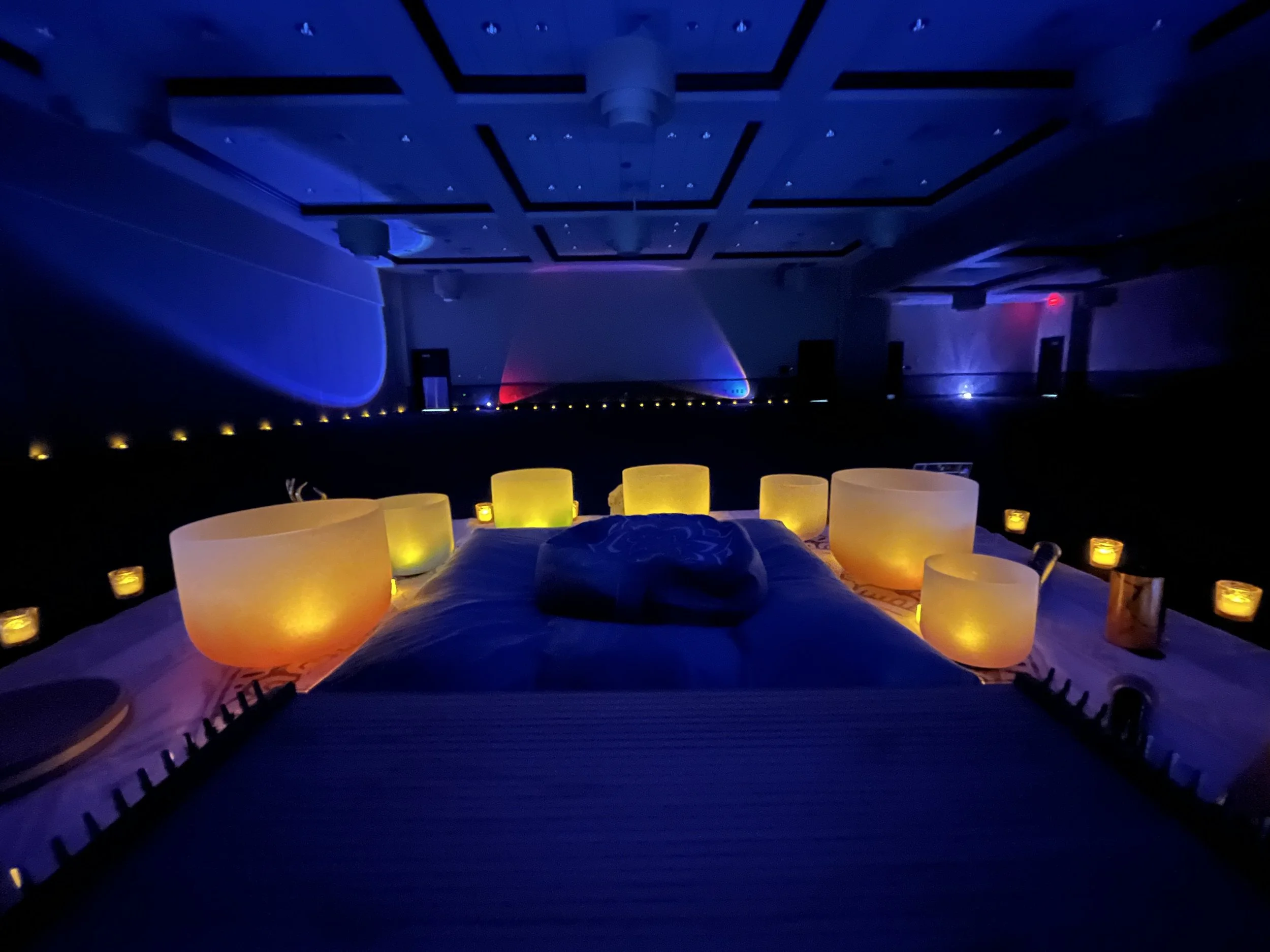 A dimly lit room with a massage or meditation setup, featuring several glowing bowls or drums arranged around a massage table and small candles along the edges.