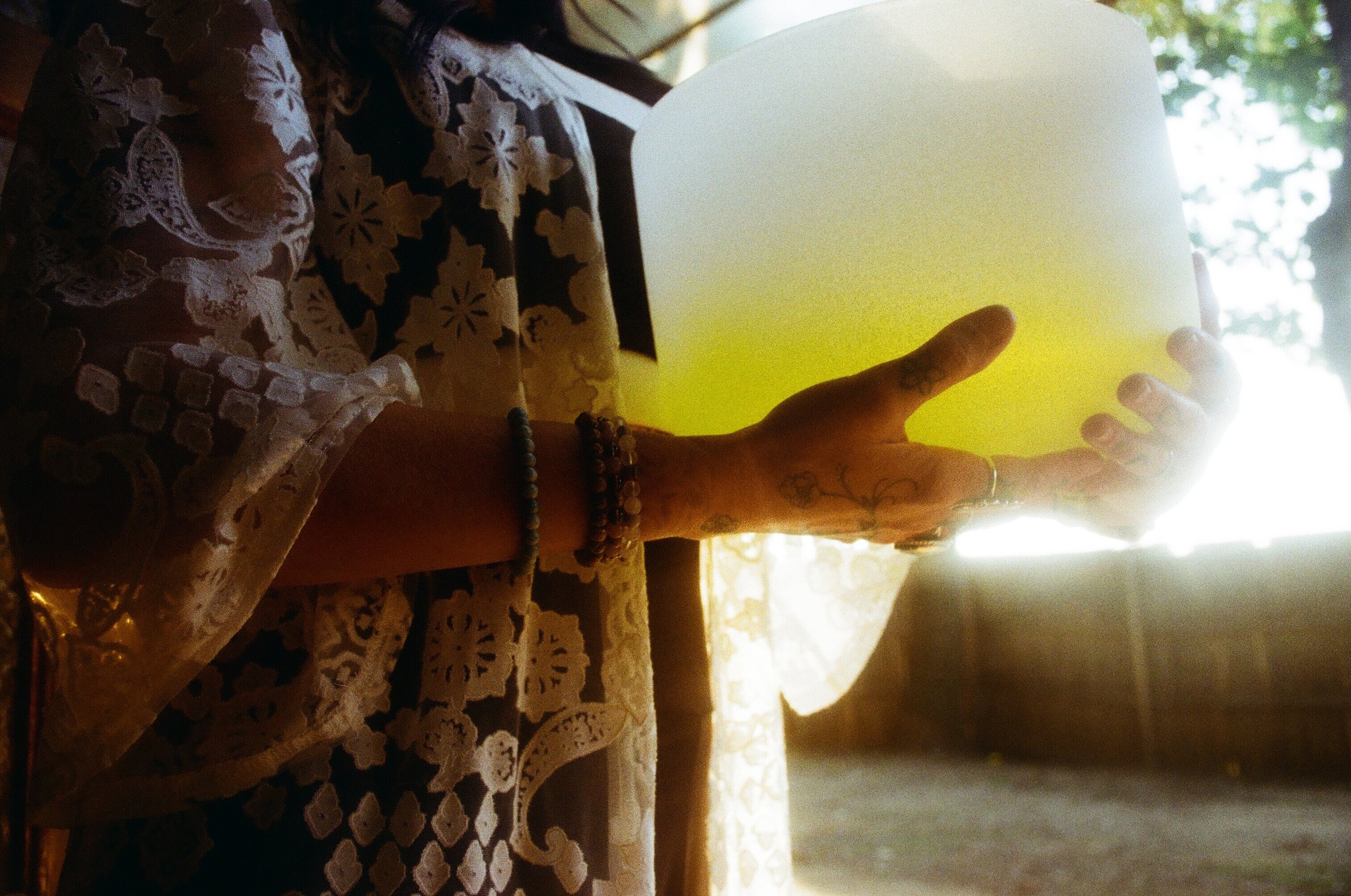 Person holding a glowing white and yellow lamp with tattoos and beaded bracelets, wearing a lace patterned garment.