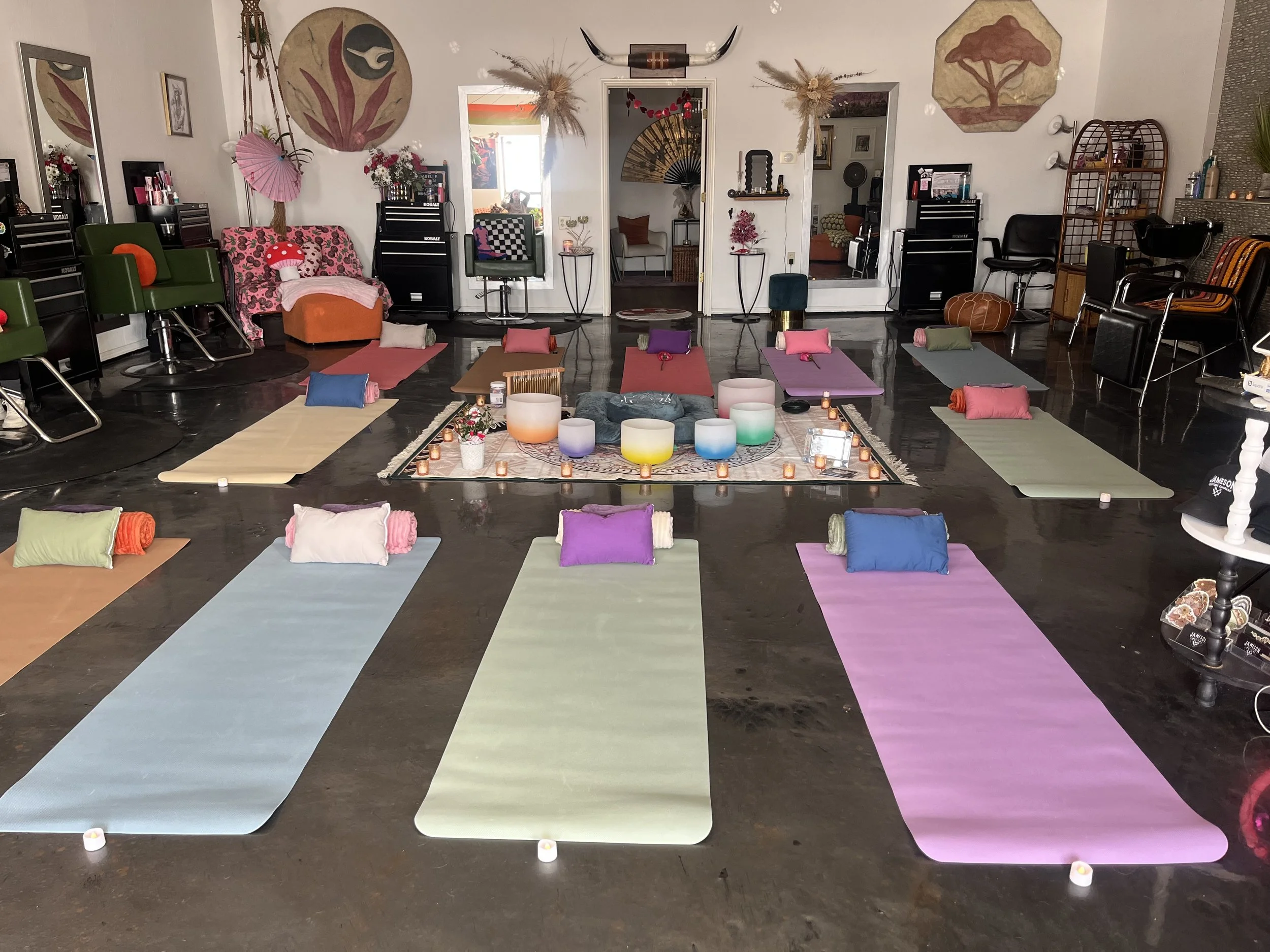 A room set up for a mindfulness or meditation class with colorful yoga mats, each with a rolled towel and a cushion. There are candles and decorative items in the center. The room has wall art, mirrors, and chairs along the walls, creating a cozy and